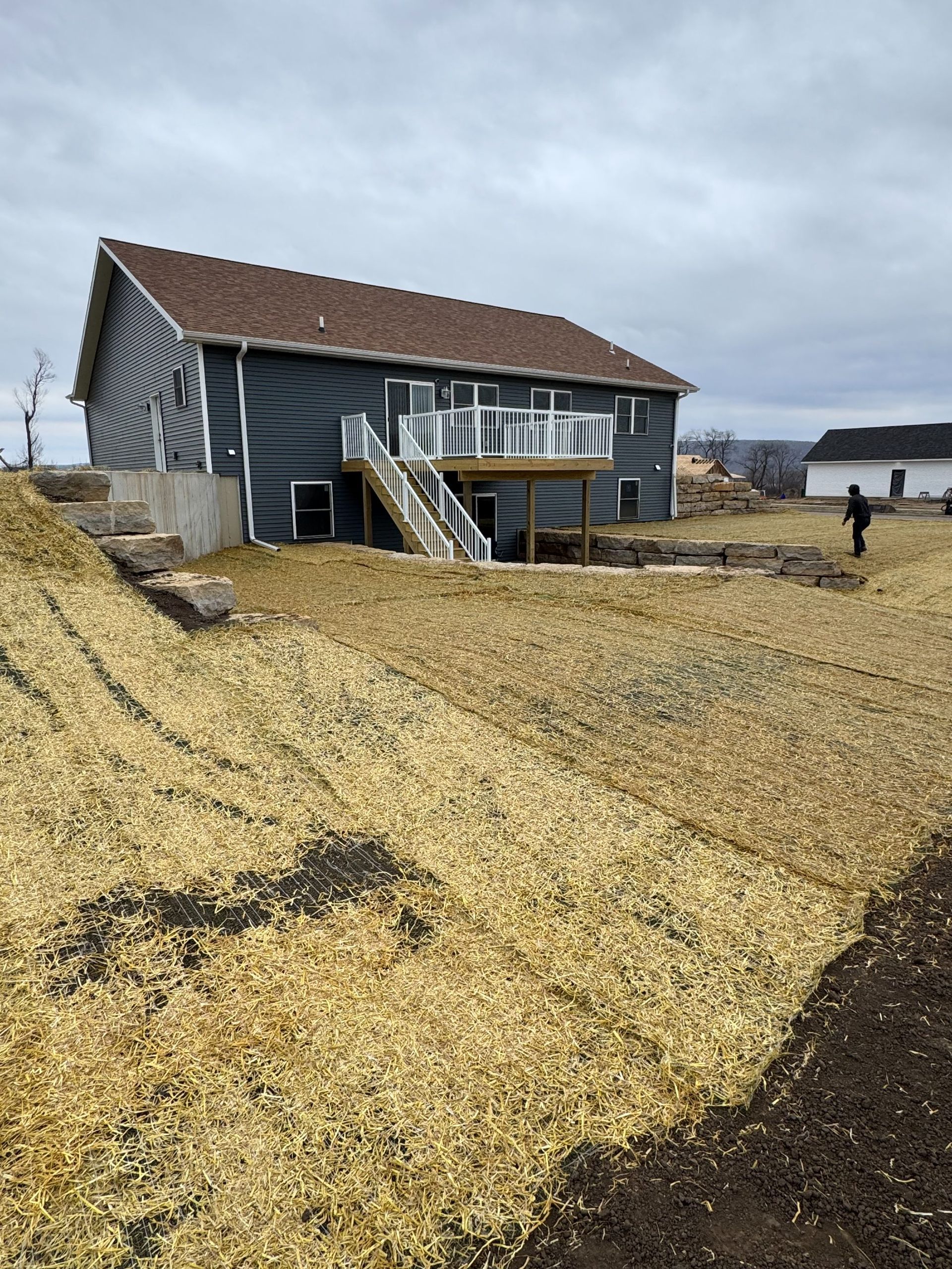 A dark blue house with a wooden deck and staircase sits on a large, straw-covered sloped yard under a cloudy sky.
