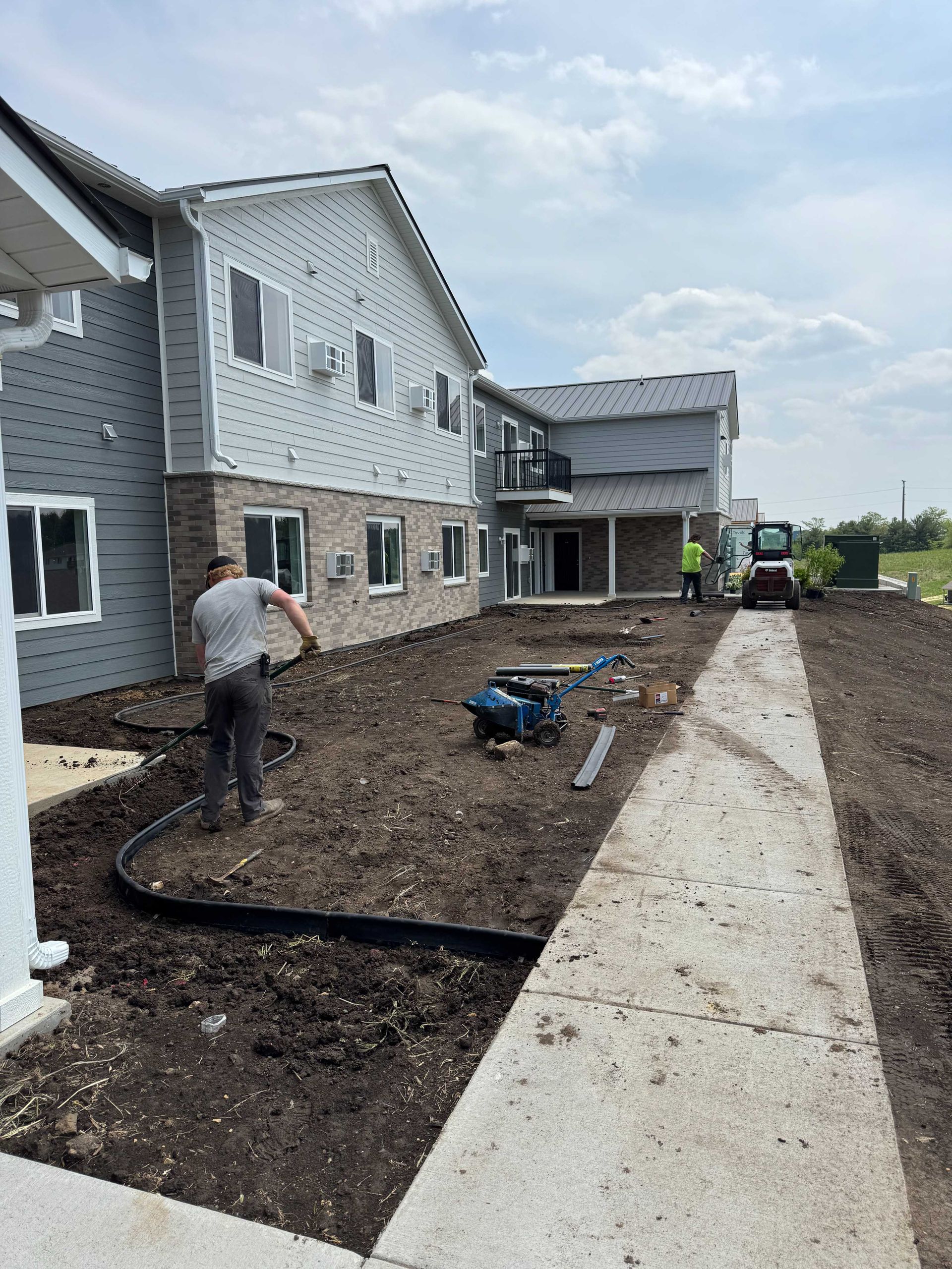 Workers install landscaping around a multi-story building under construction with a grey exterior and stone foundation.