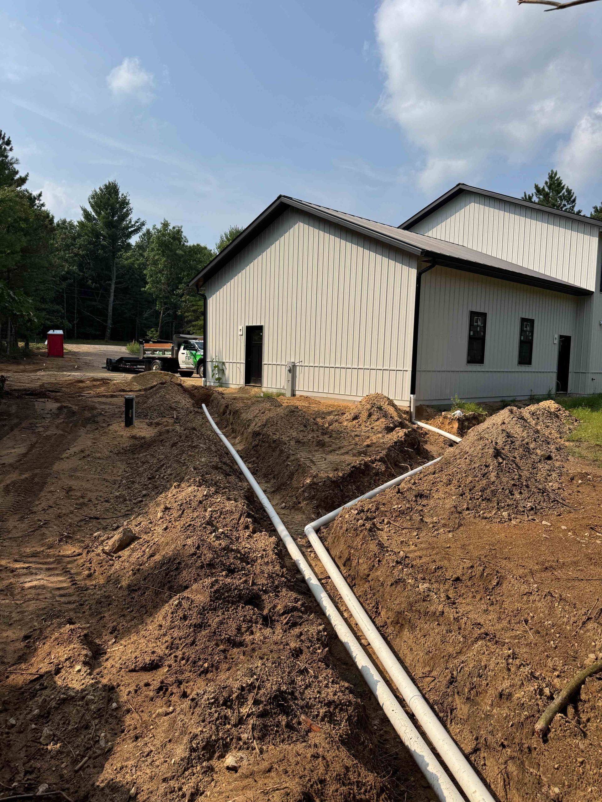 Two white PVC pipes lie in an open dirt trench leading toward the side of a modern white metal building.