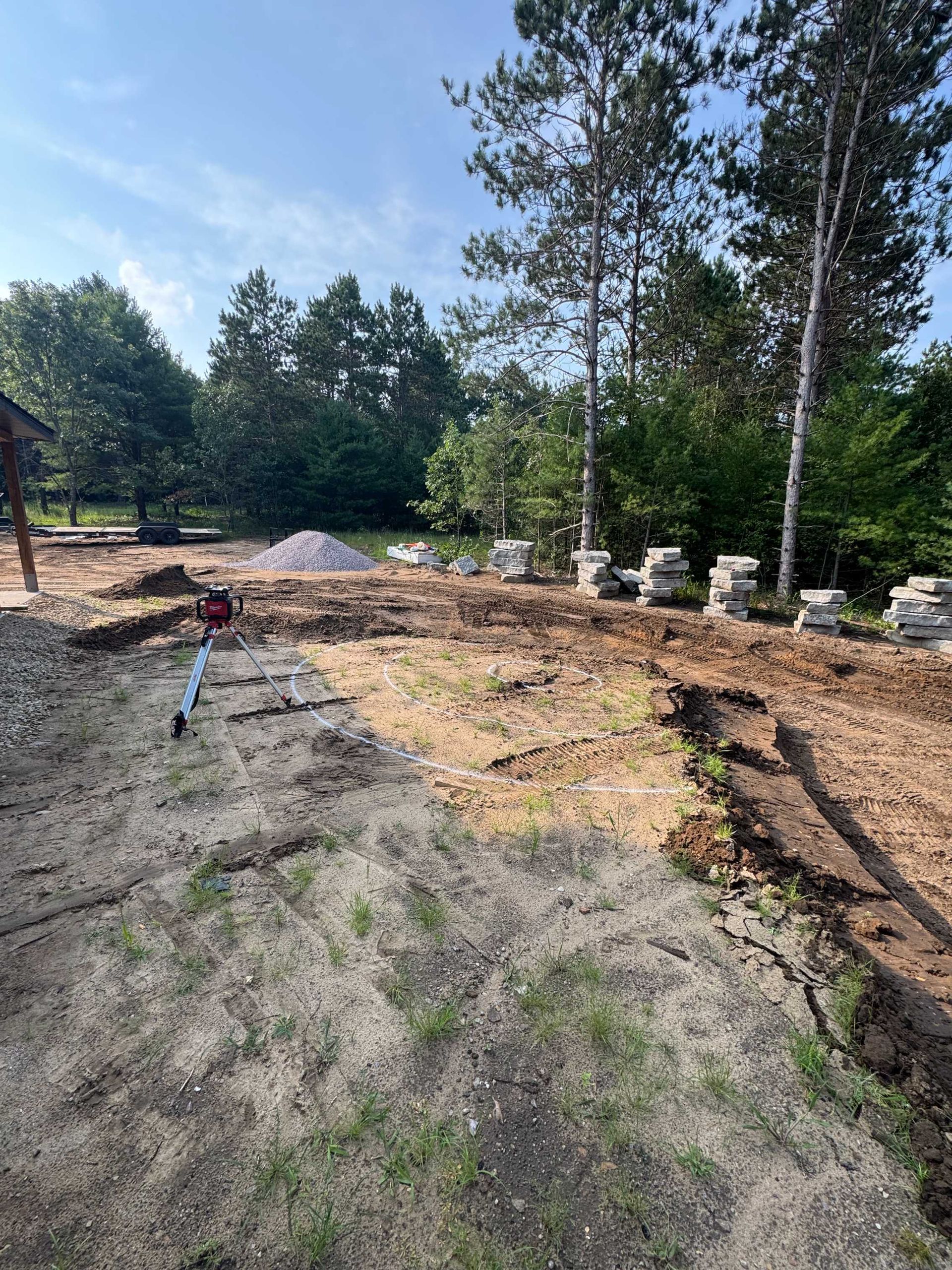 A rural landscape showing a construction site with a freshly dug trench, scattered stone blocks, and a pile of gravel.