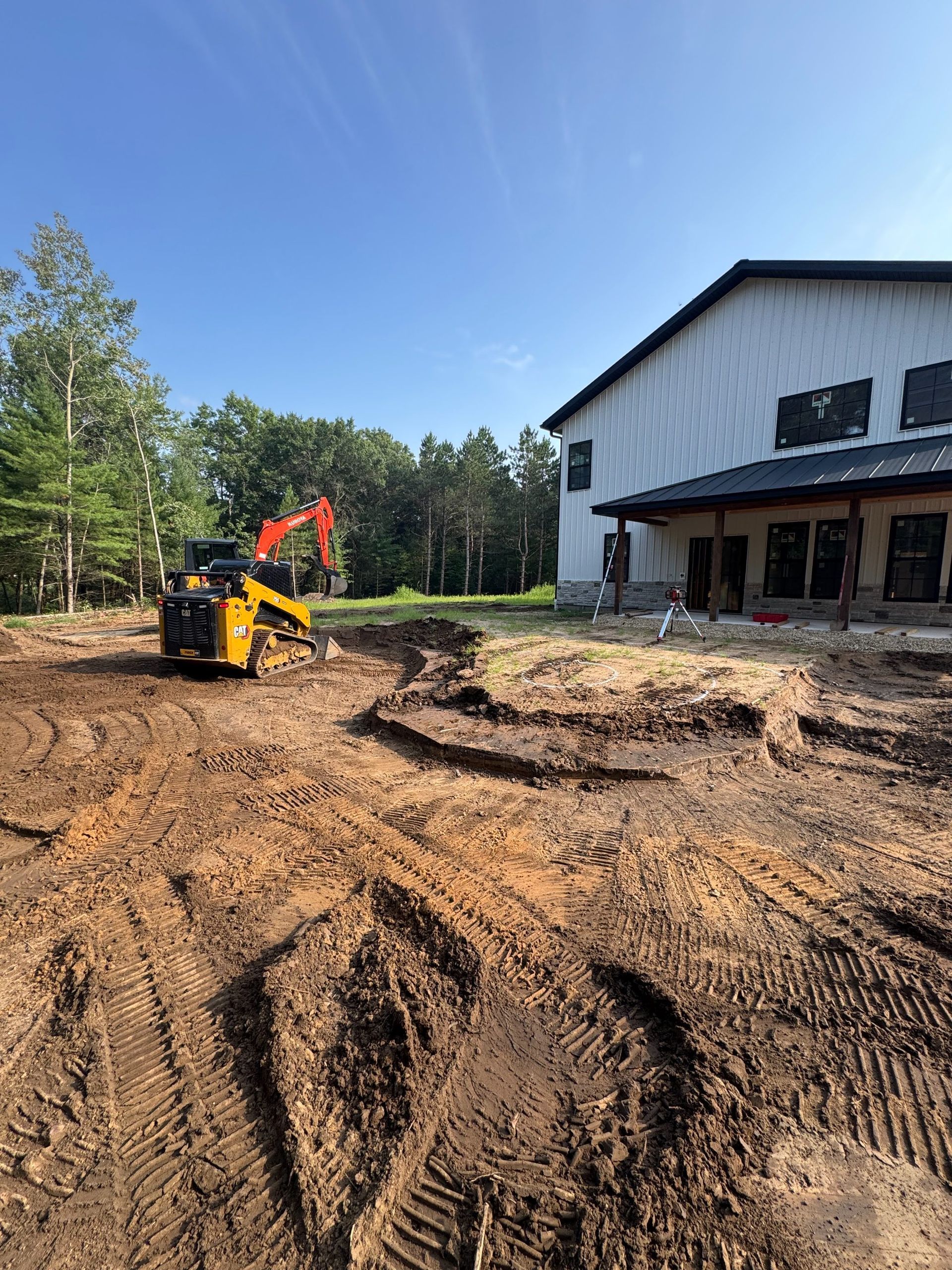 A bright yellow skid steer excavator parked on a muddy construction site next to a white building with black accents.