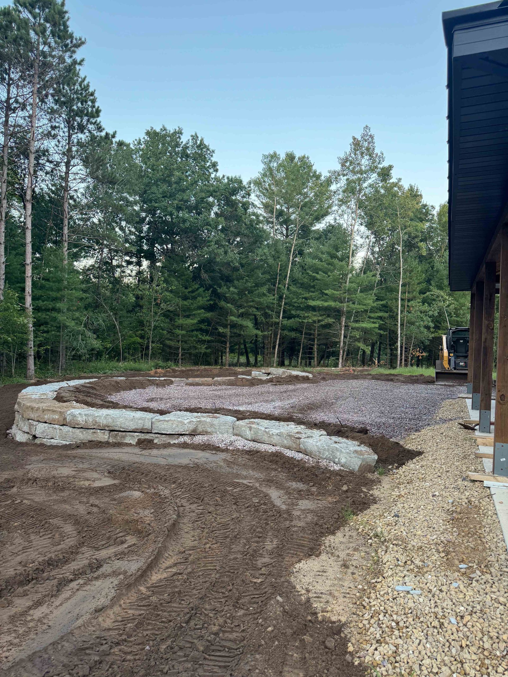 A newly constructed stone retaining wall bordering a gravel patio area next to a house, with a wooded background.