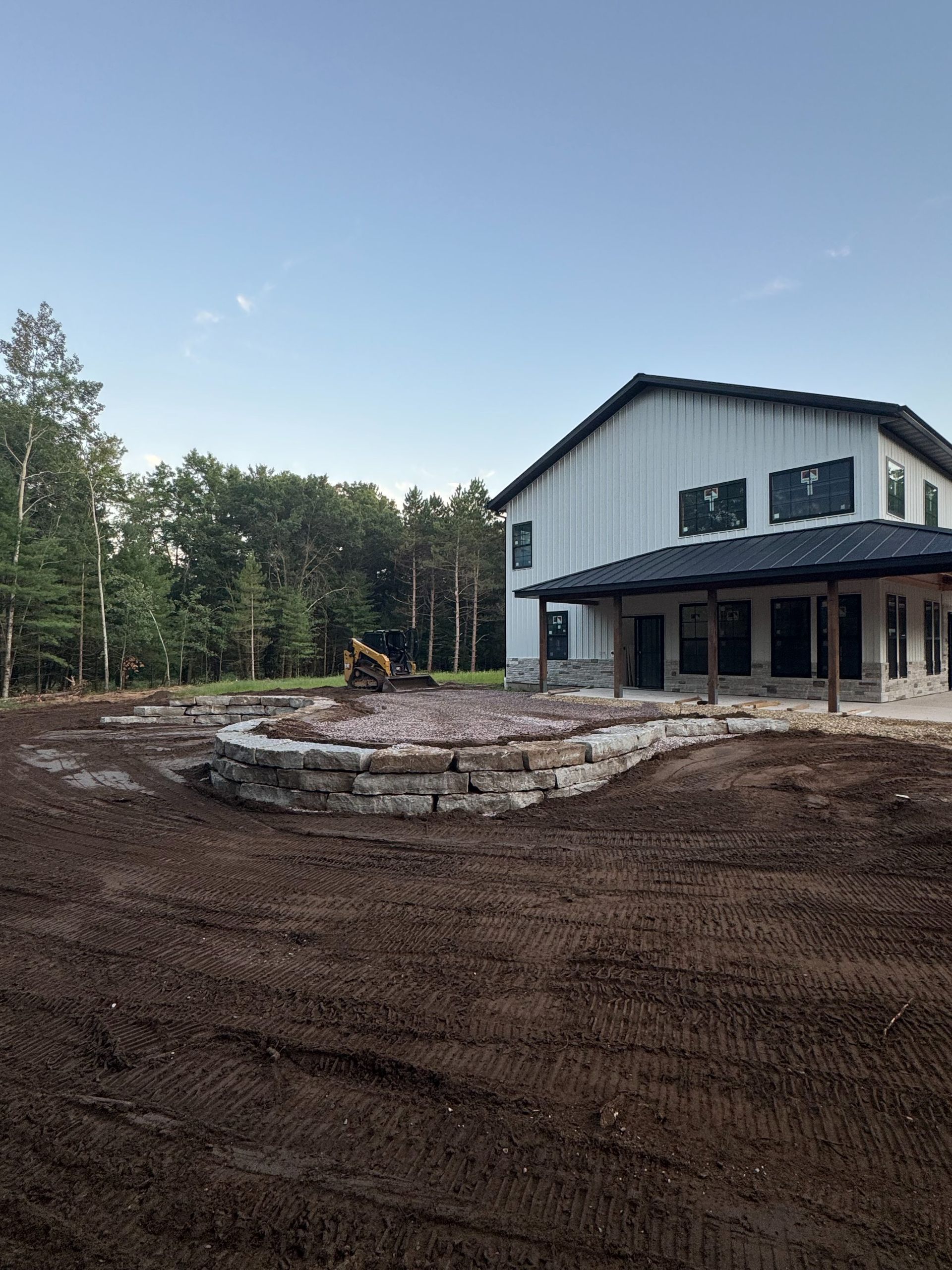 A modern, light-colored barn-style building sits behind a freshly laid curved stone retaining wall and dirt grounds.