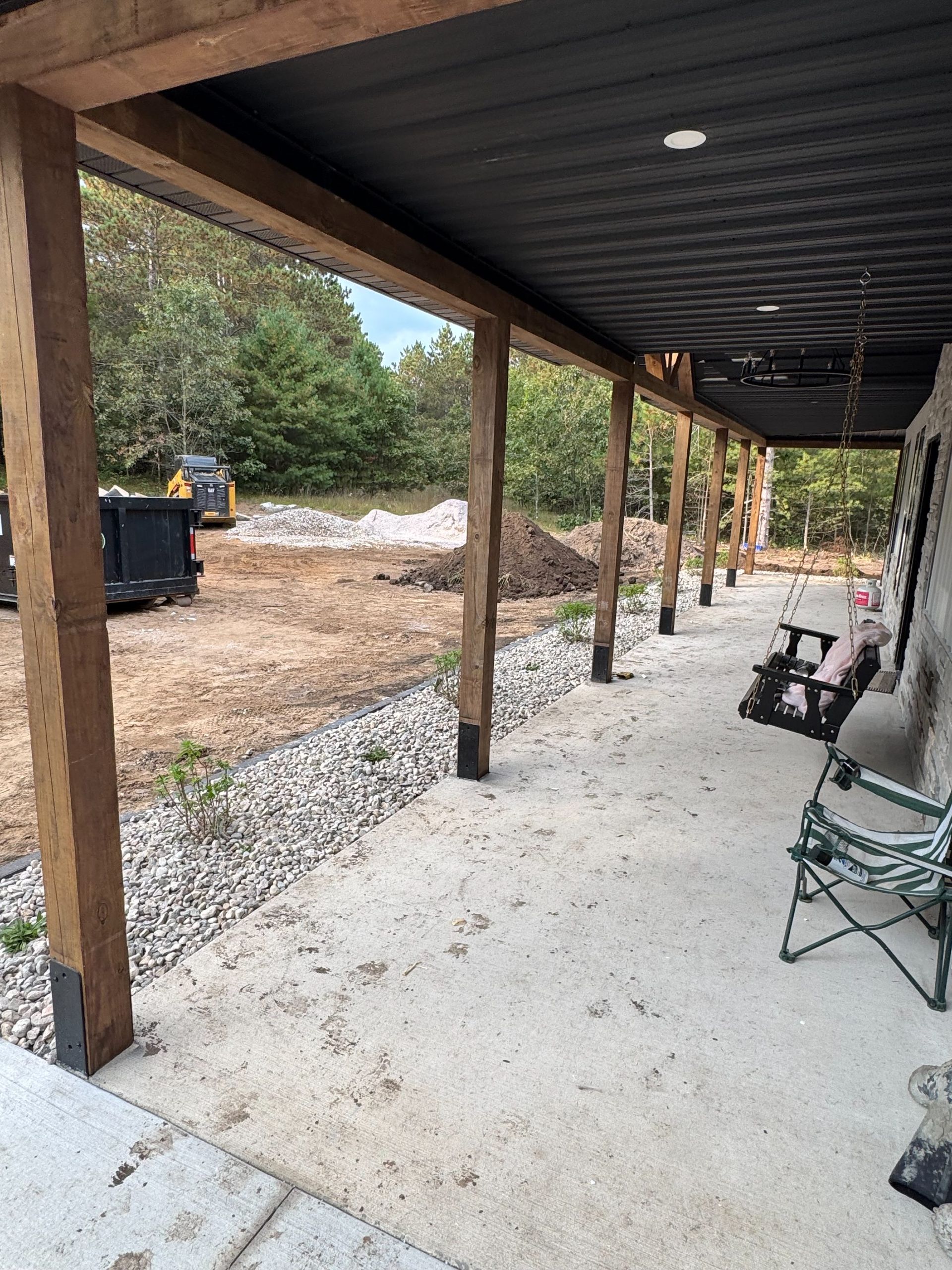 A covered concrete porch with wooden support posts, overlooking a construction site with a skip and a pile of dirt.
