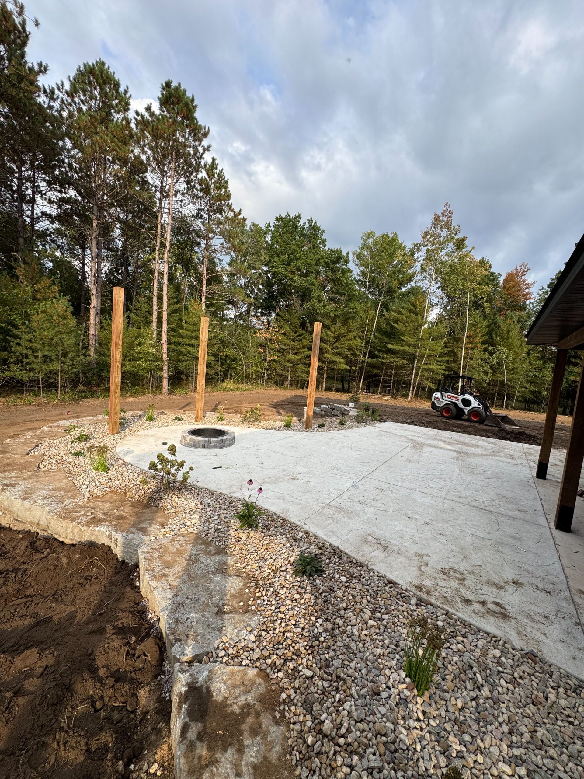 A light gravel patio area with a fire pit, bordered by stone and vertical wooden posts against a backdrop of trees.