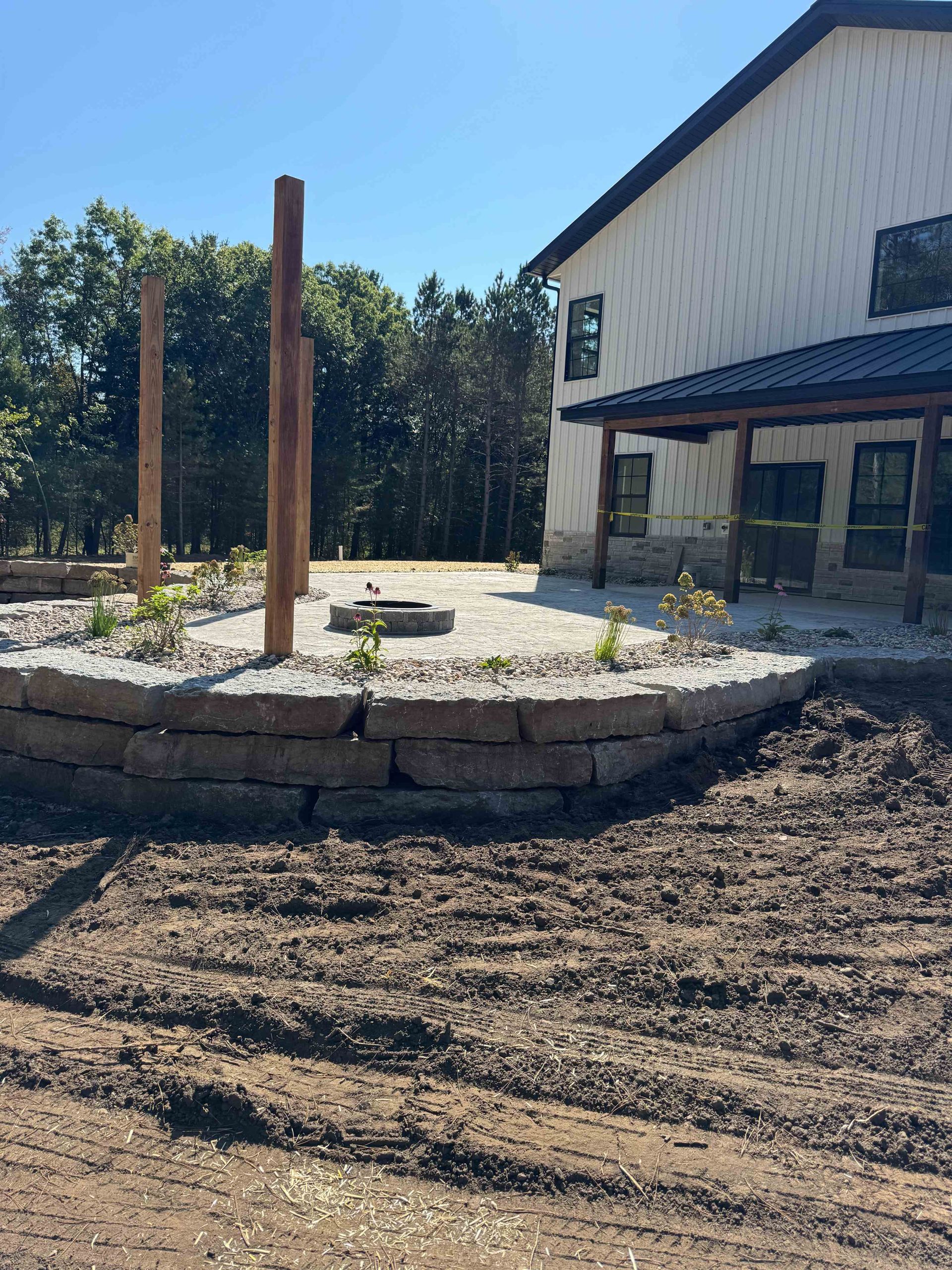 A stone patio with a fire pit and wooden posts under construction next to a modern building with a metal roof.