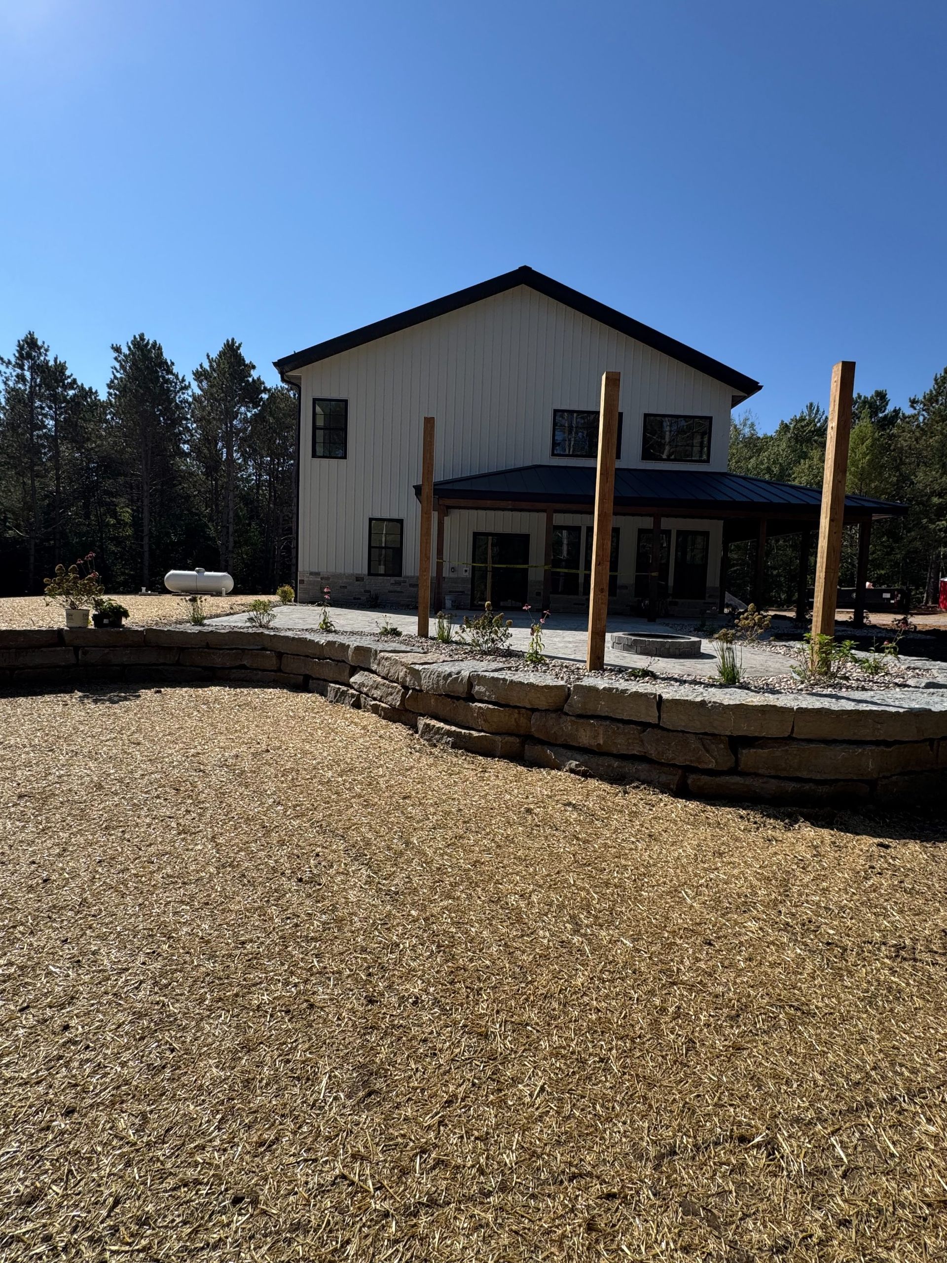 A two-story white house with a patio, wooden support posts, and a stone retaining wall in a wood-chipped yard.