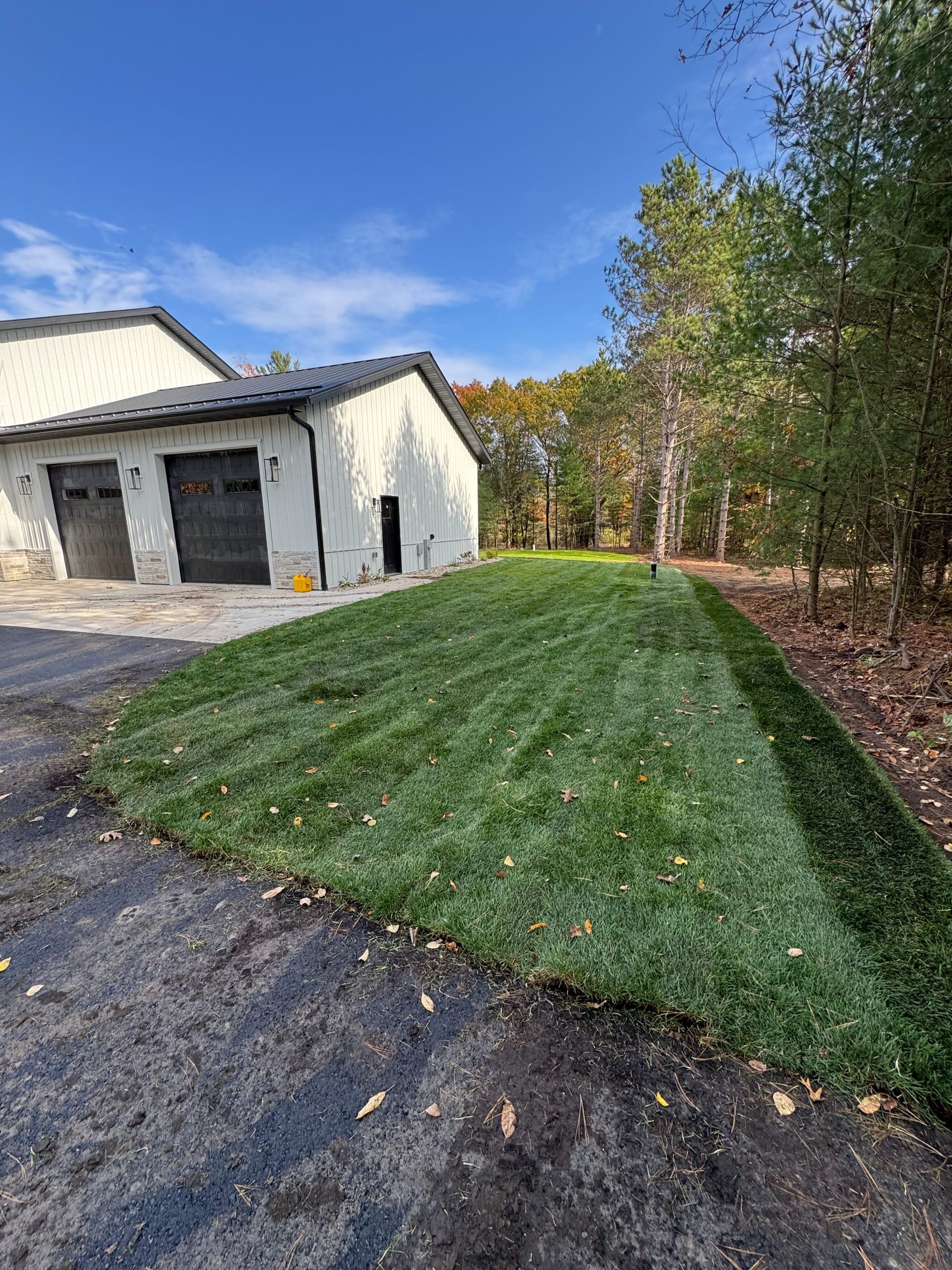 Freshly laid green sod borders a light-colored building with two dark garage doors next to a tree line under a blue sky.