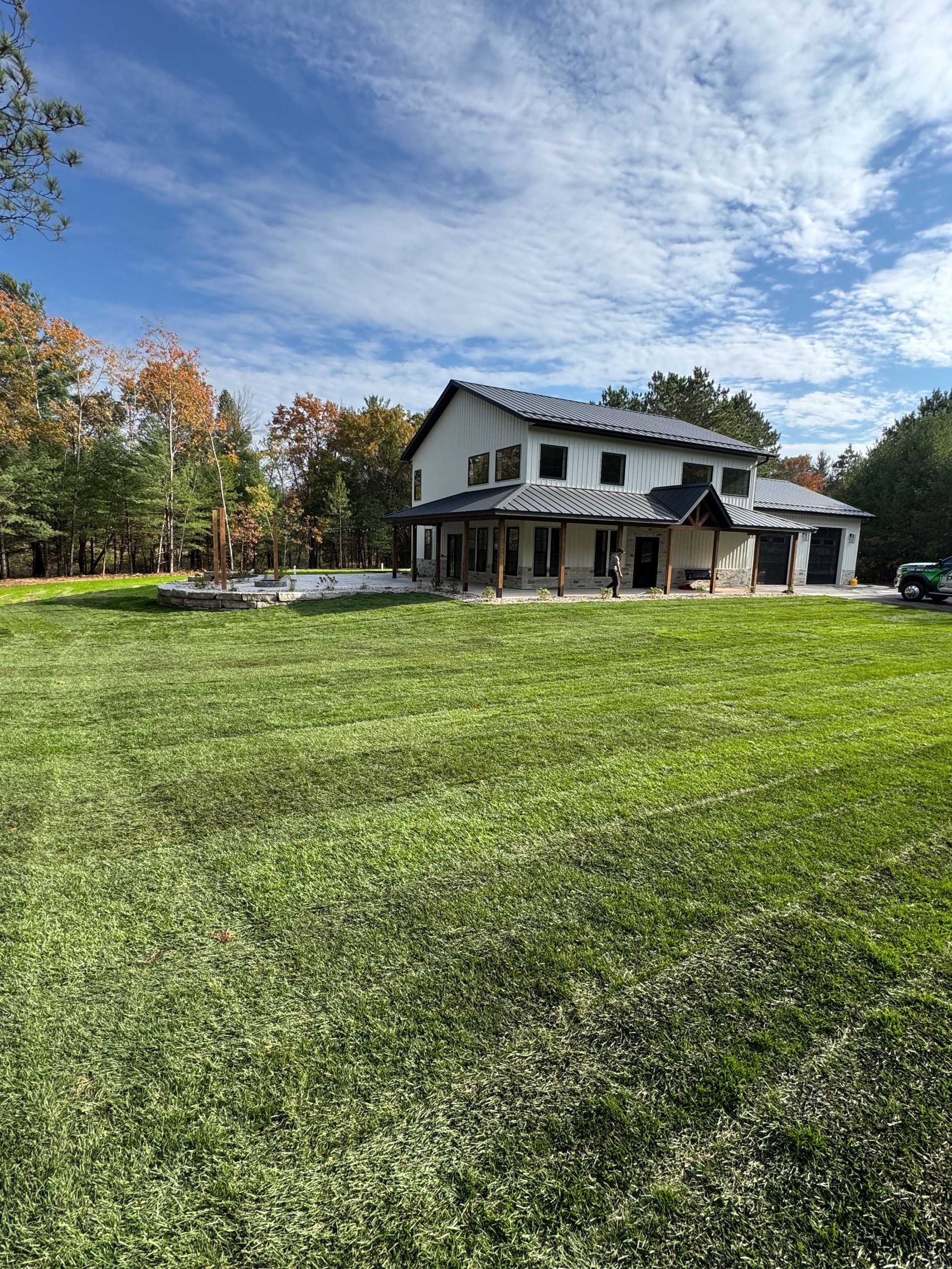 A two-story house under construction stands in a large, sunny field surrounded by trees with autumn leaves.