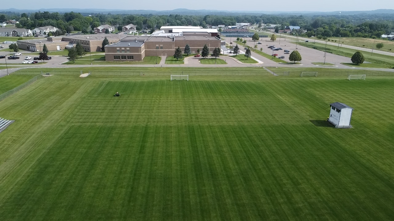 An aerial view of a grass sports field with a small white shed, adjacent to a large school building and parking lot.