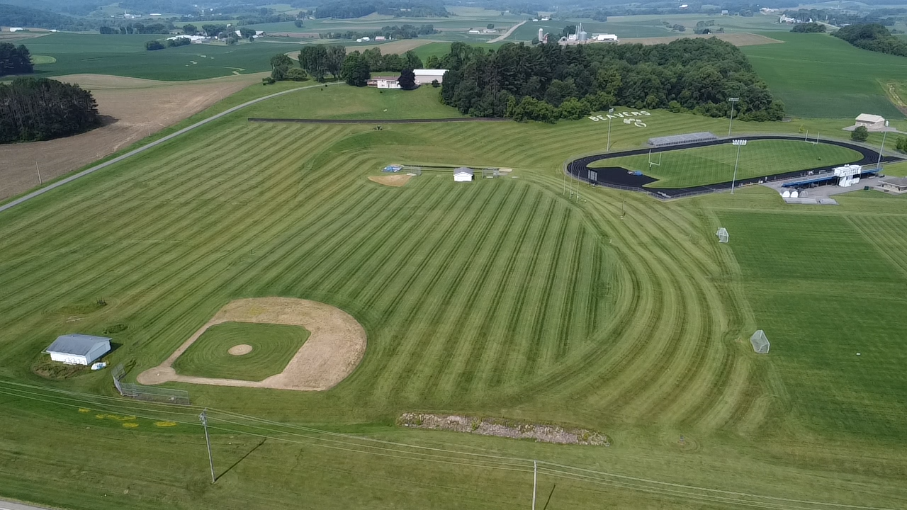An aerial view of a baseball field and an oval running track surrounded by green farmland and trees.