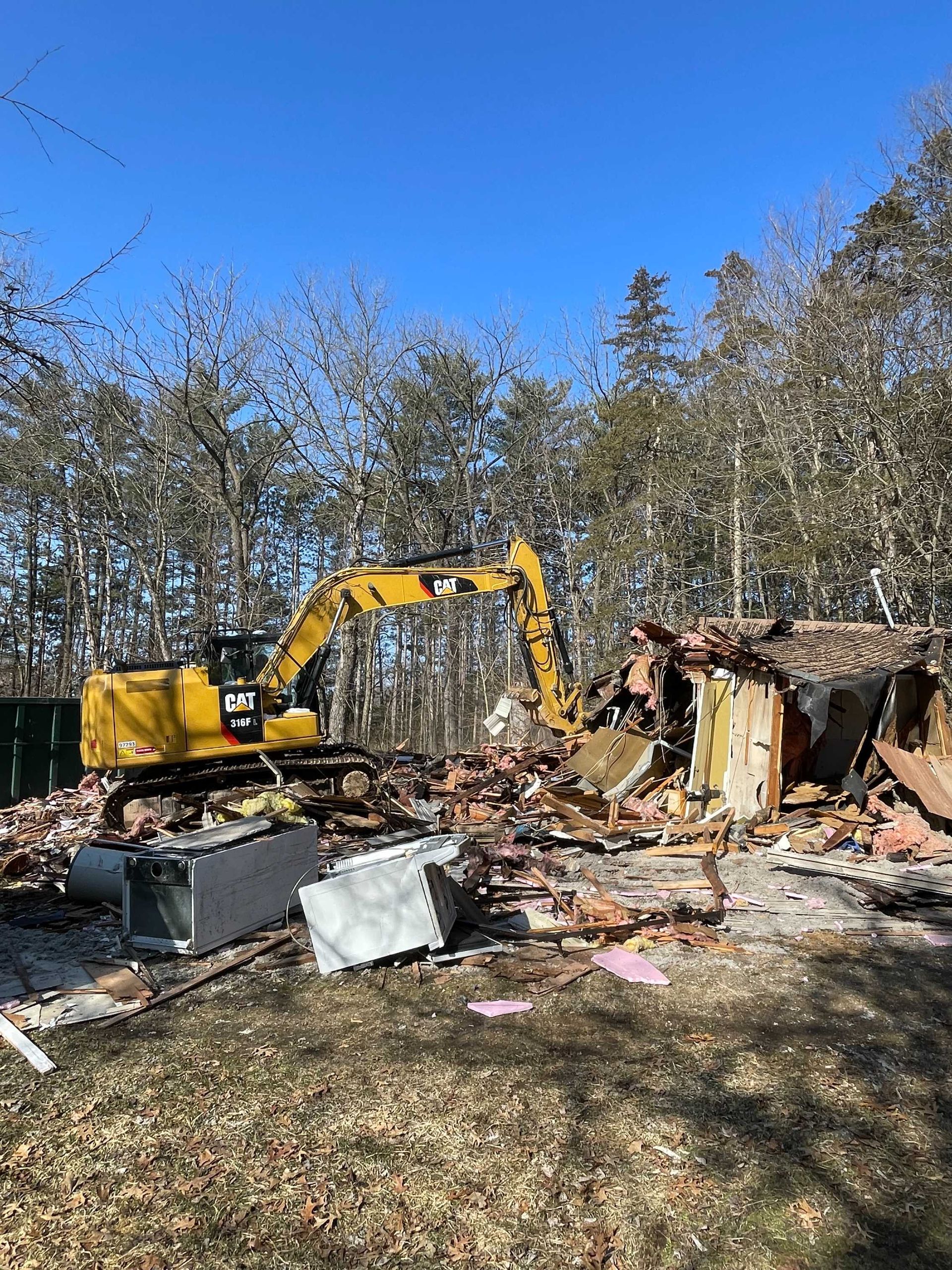 A yellow Caterpillar excavator demolishing a structure in a wooded area on a sunny day.