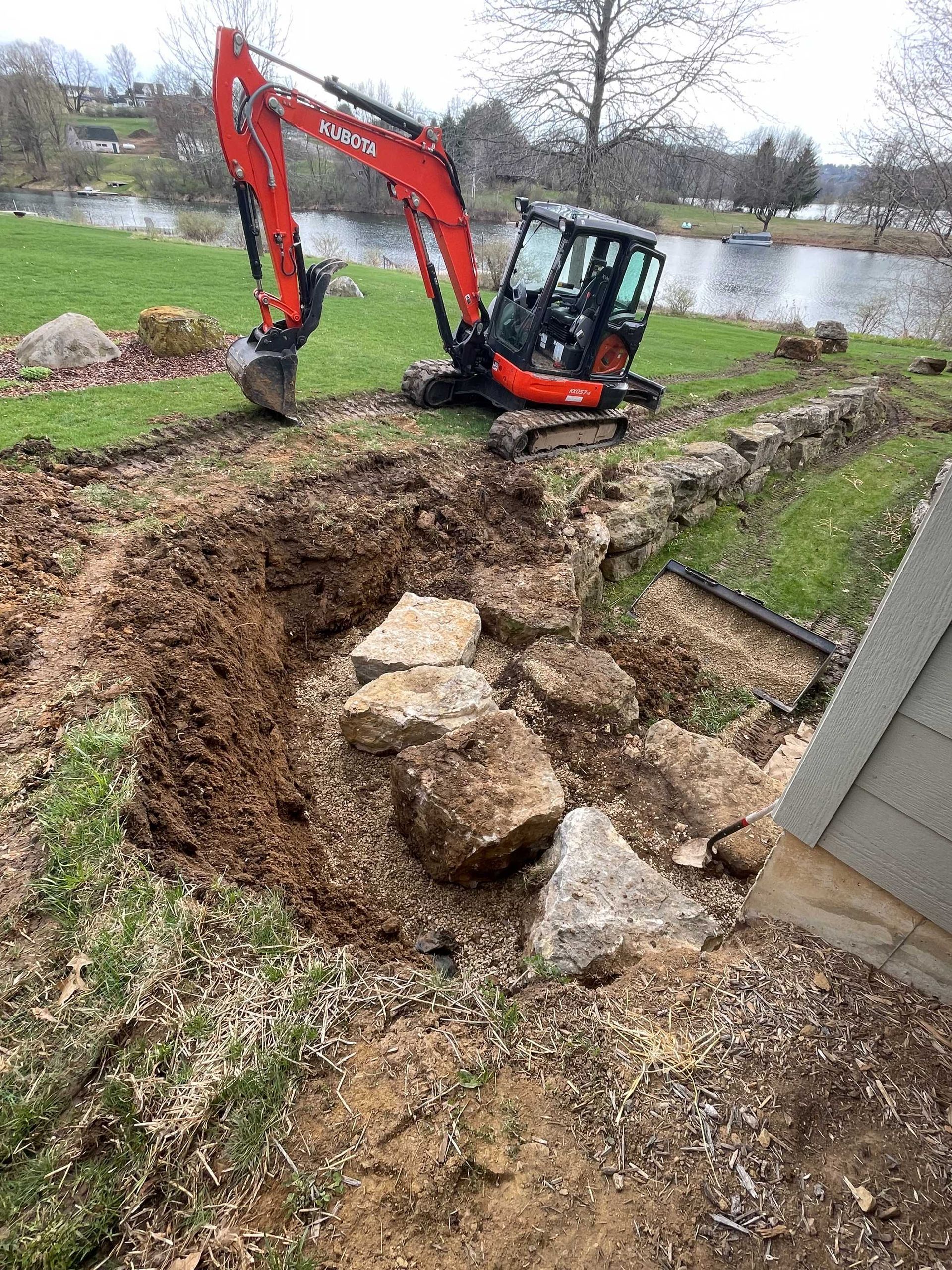 A red excavator parked by a lake, working on a landscaping project with large rocks in an excavated dirt trench.