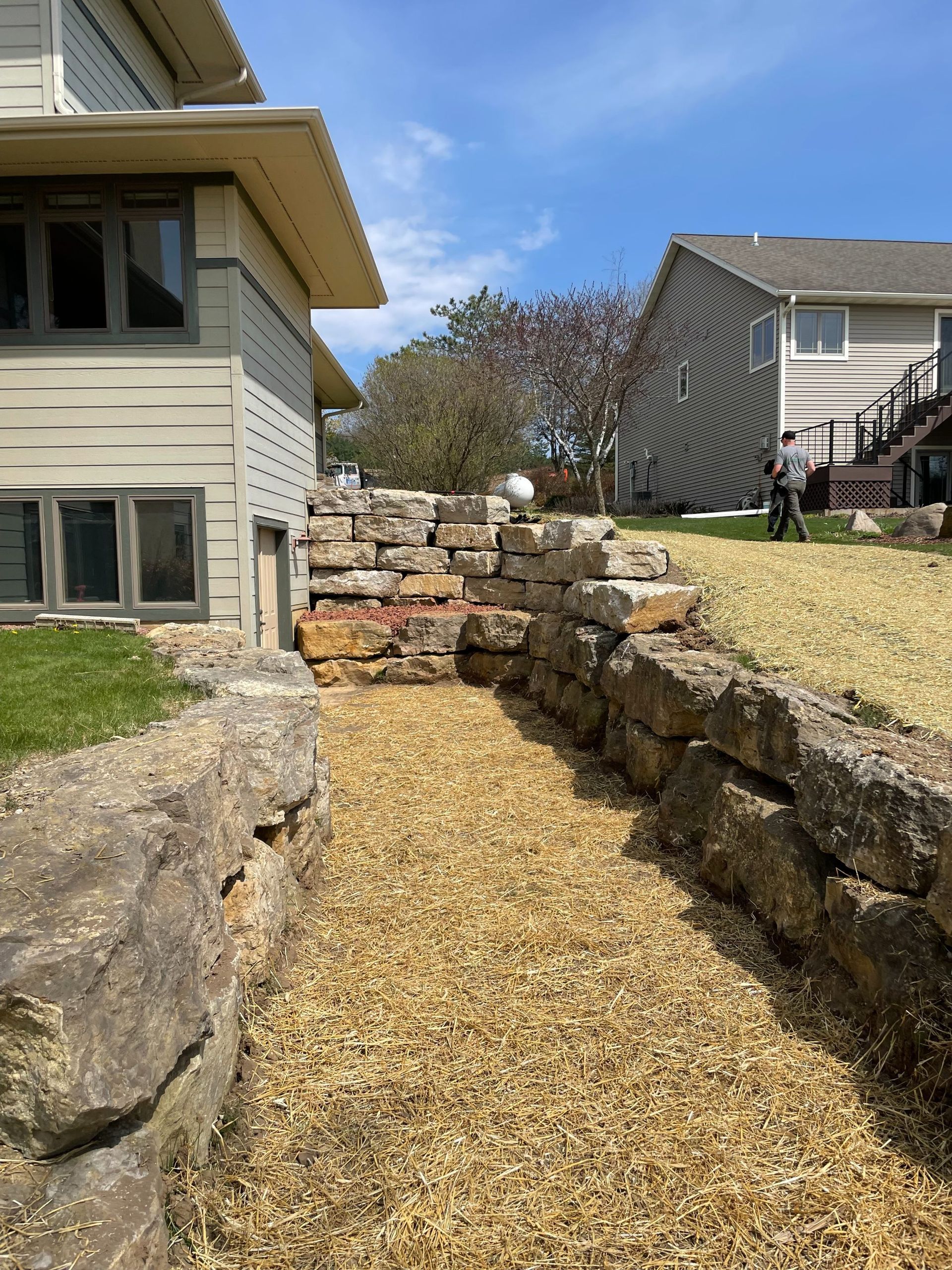 A stone retaining wall flanks a gravel path leading between a house and a grassy yard under a blue sky.