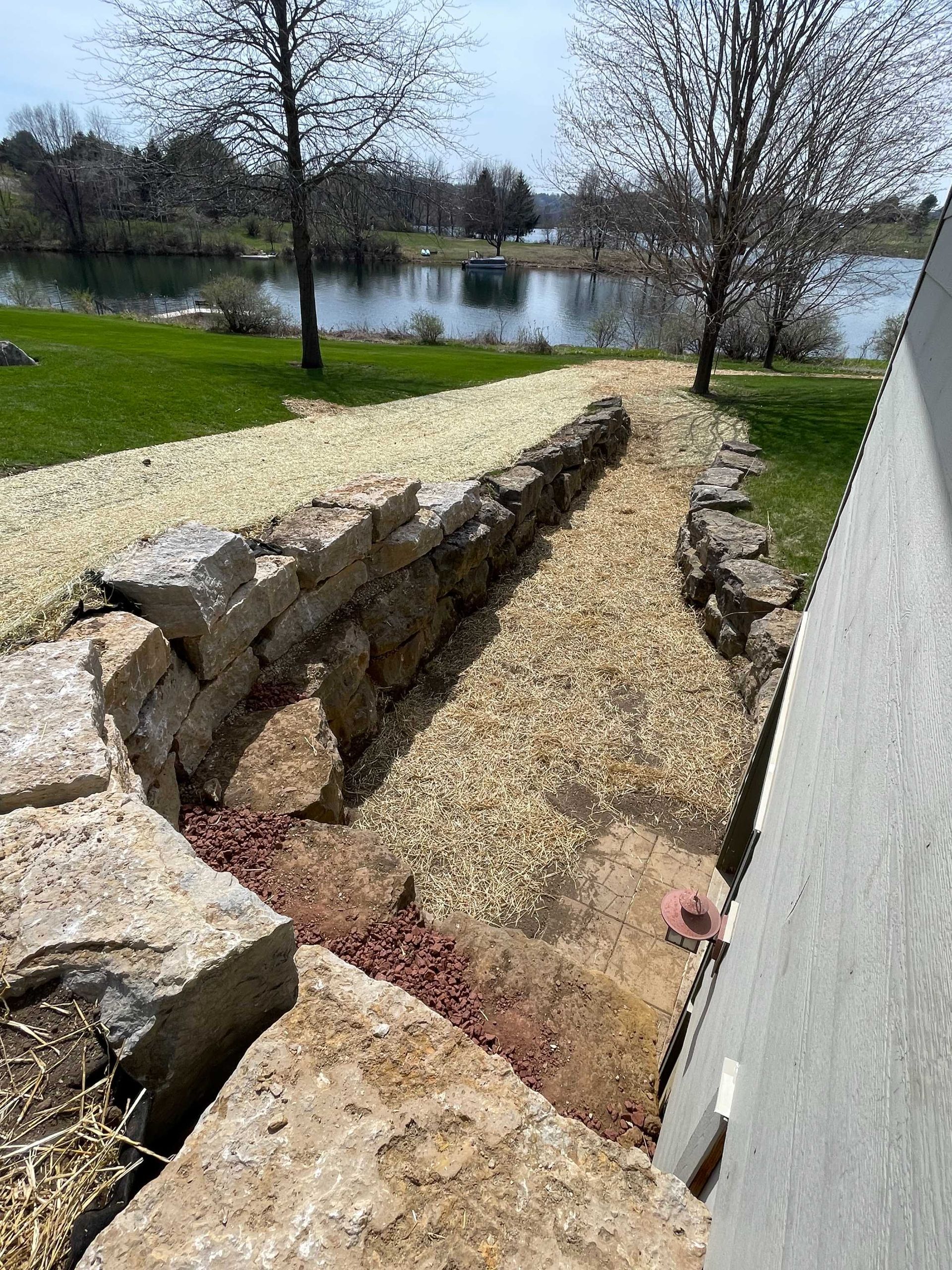A stone retaining wall lining a gravel path beside a building, with a grassy yard and lake in the background.