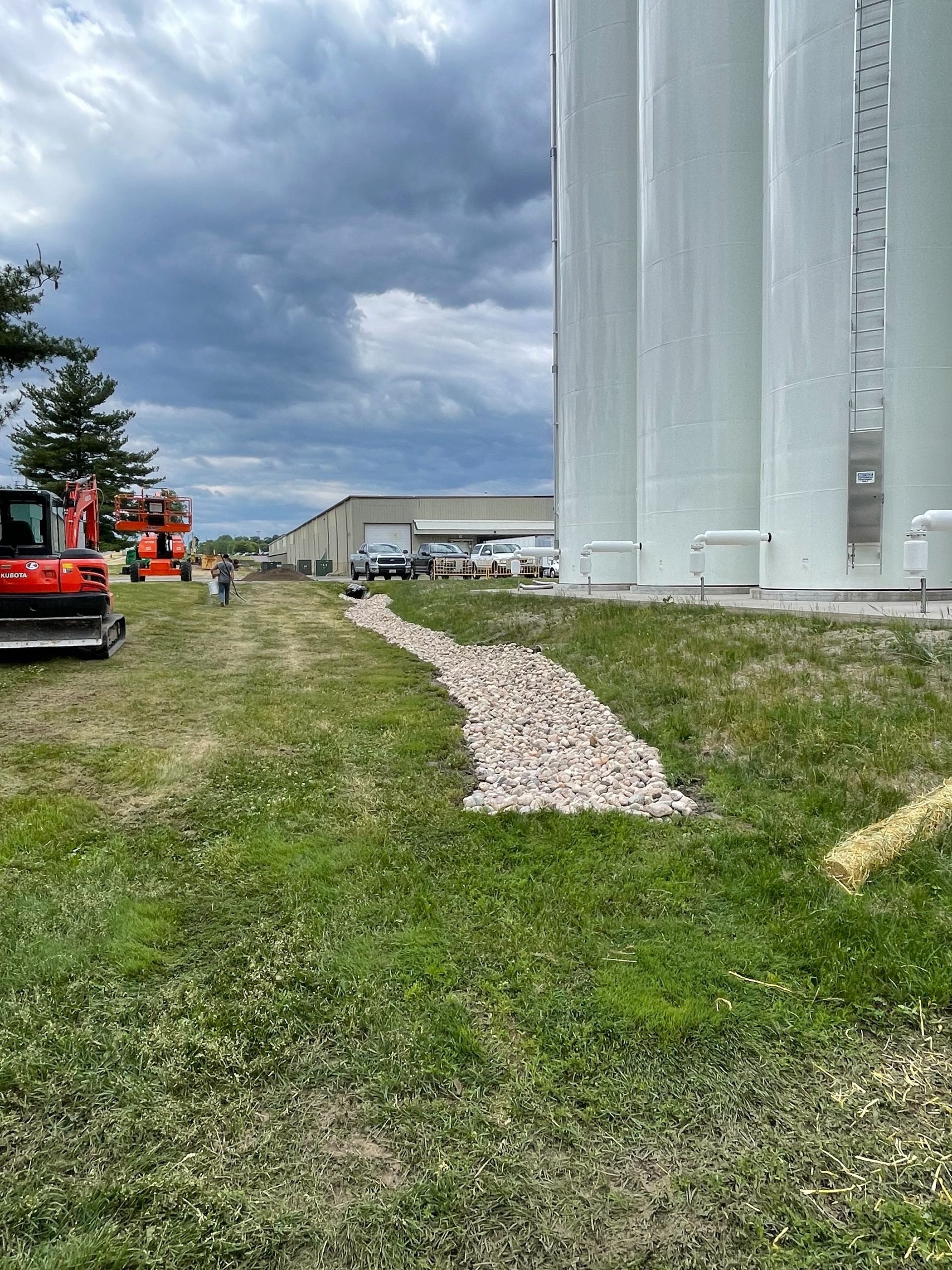A gravel drainage path runs along a row of tall white storage silos on a grassy site under a cloudy sky.