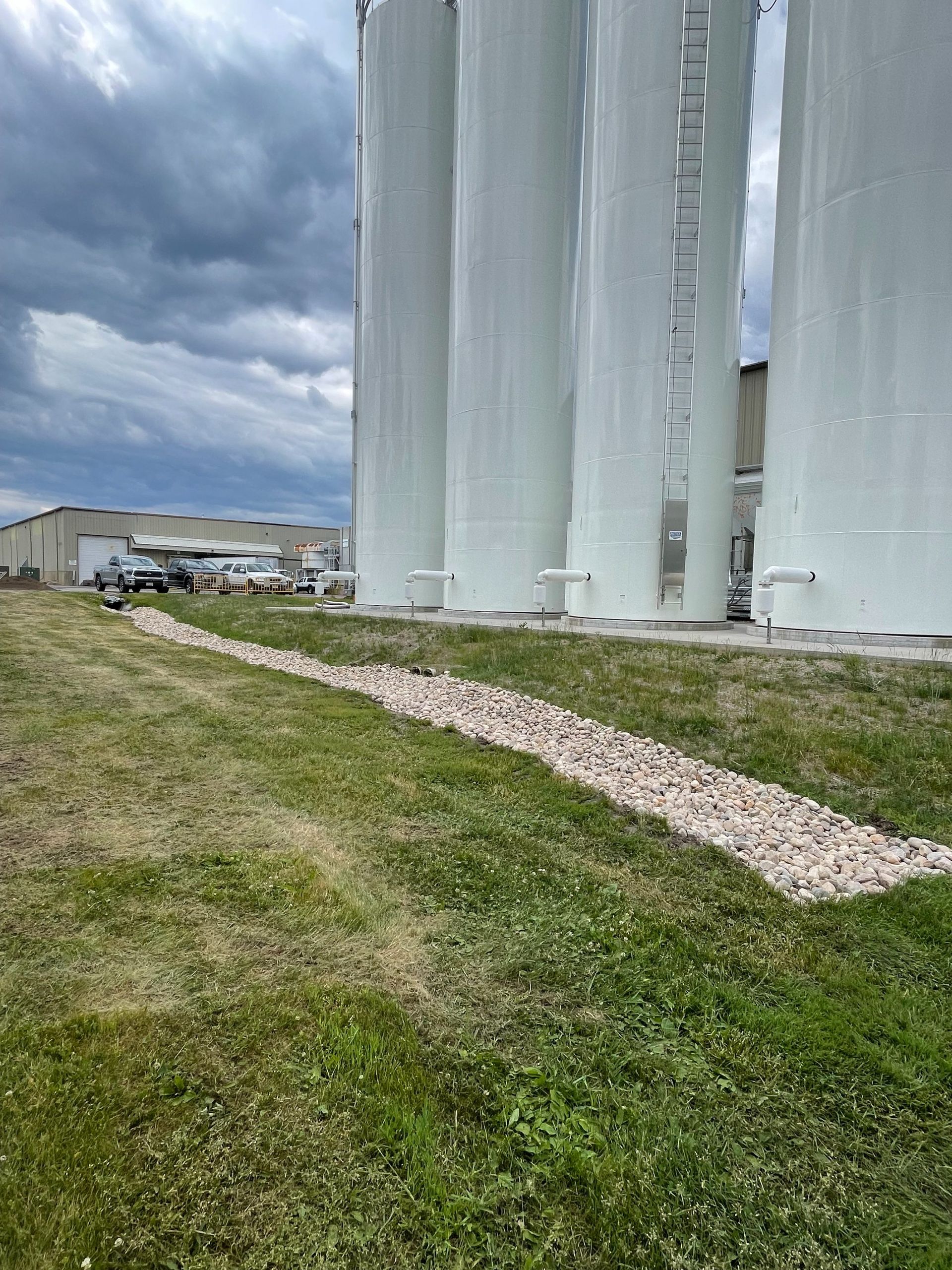 Four white industrial silos stand outdoors on a concrete base next to a gravel path and grassy field under a cloudy sky.