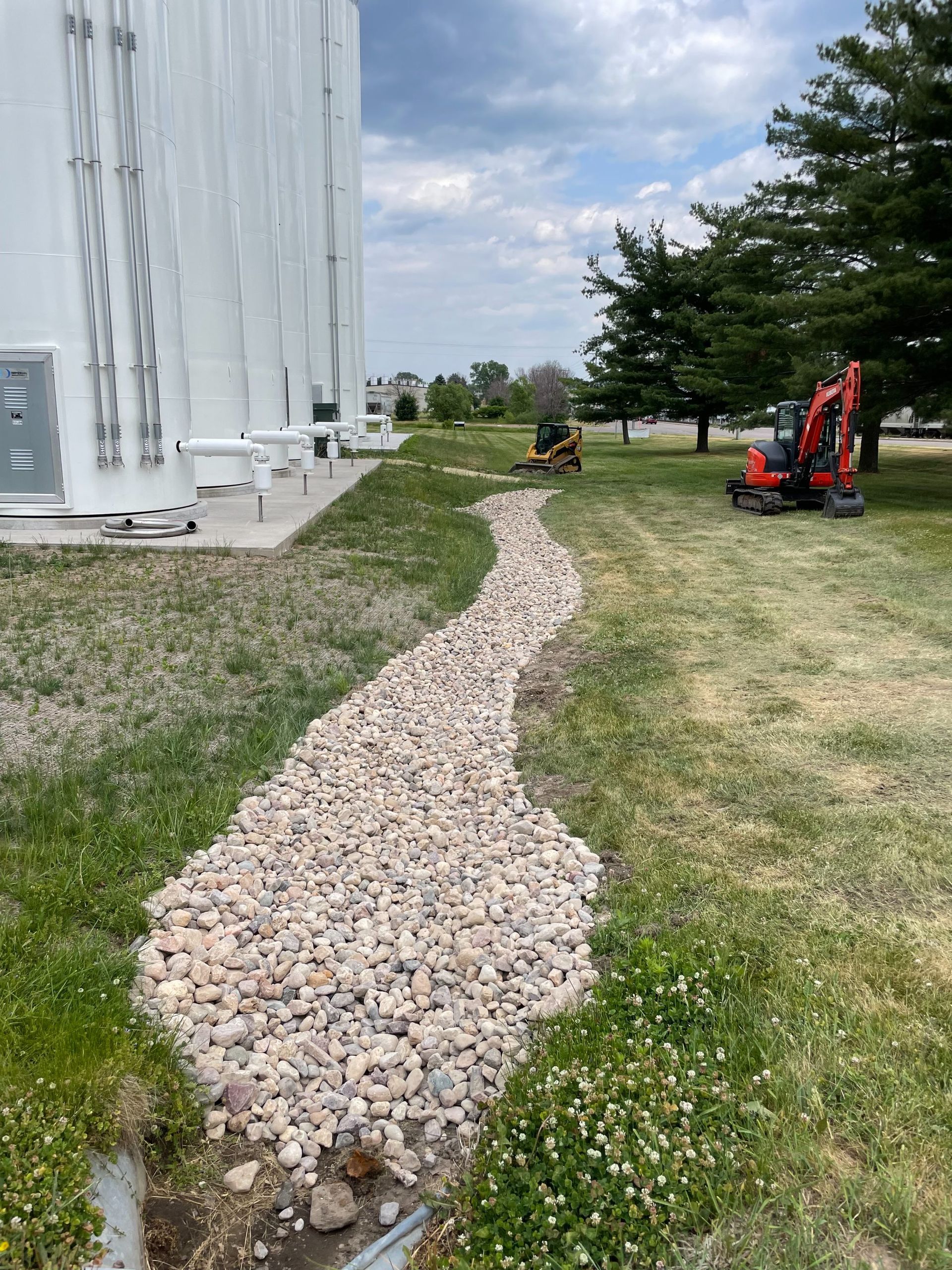 A stone-lined drainage swale runs along a white industrial tank on a grassy lawn with a small excavator in the background.