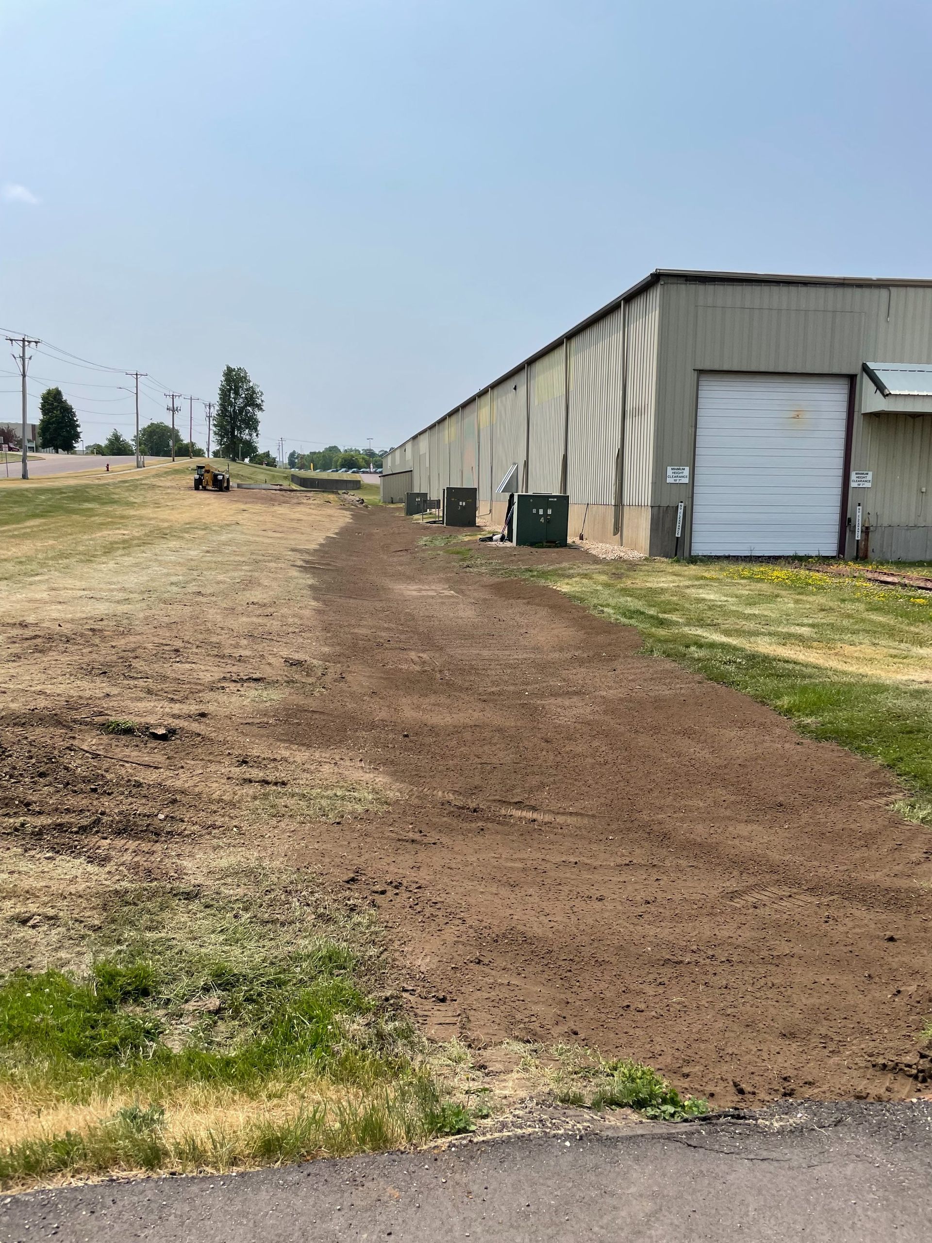 A dirt path runs alongside a long, industrial metal building with several dark storage units and a roll-up door.