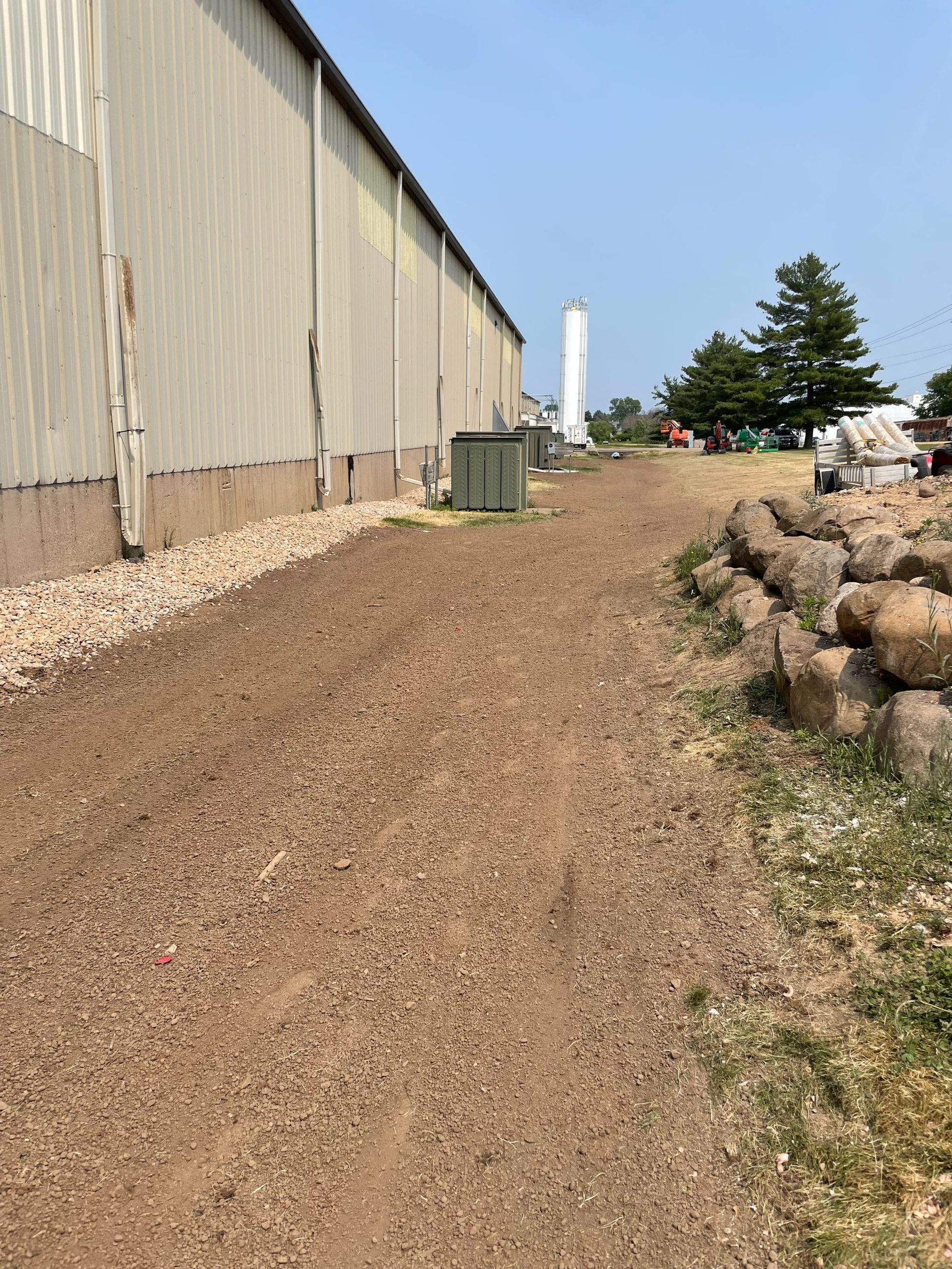 A gravel path runs beside a tall metal building wall, with a rock wall on the right and a distant water tower.