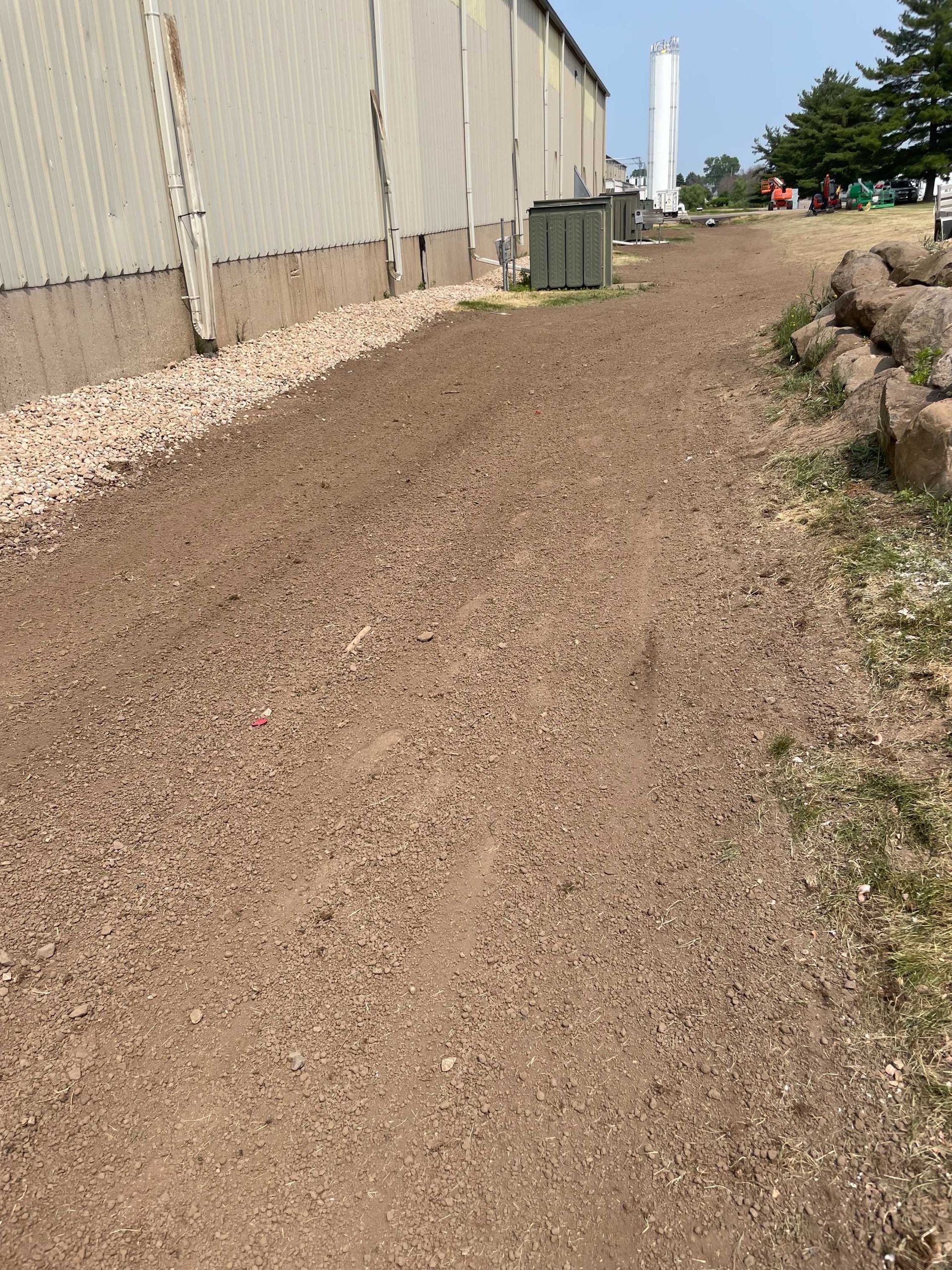 A dirt path runs alongside a metal building, bordered by light-colored stones on one side and a large rock wall on the other.