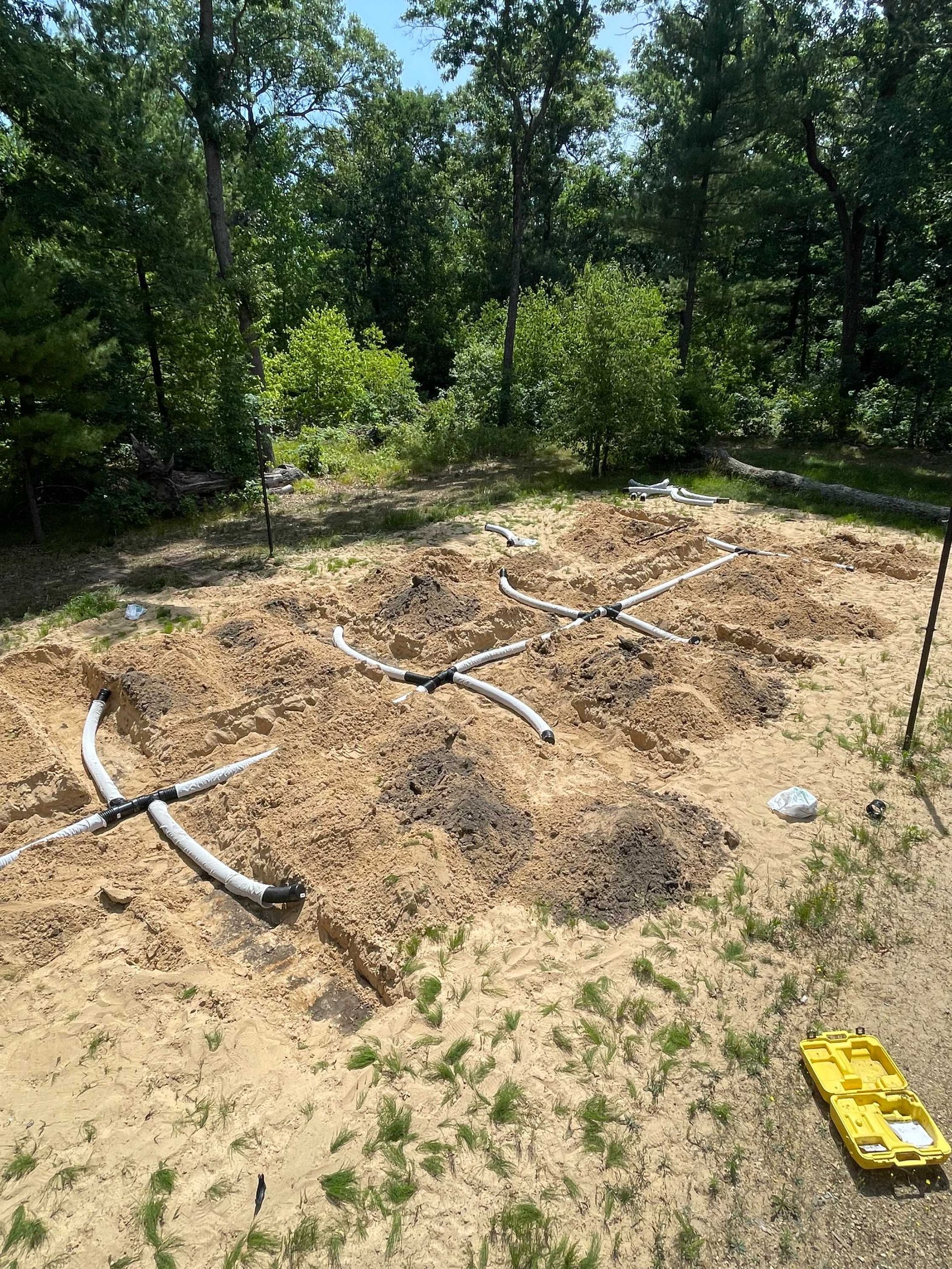 A garden plot with mounded soil and white irrigation pipes laid out on the ground, surrounded by trees.