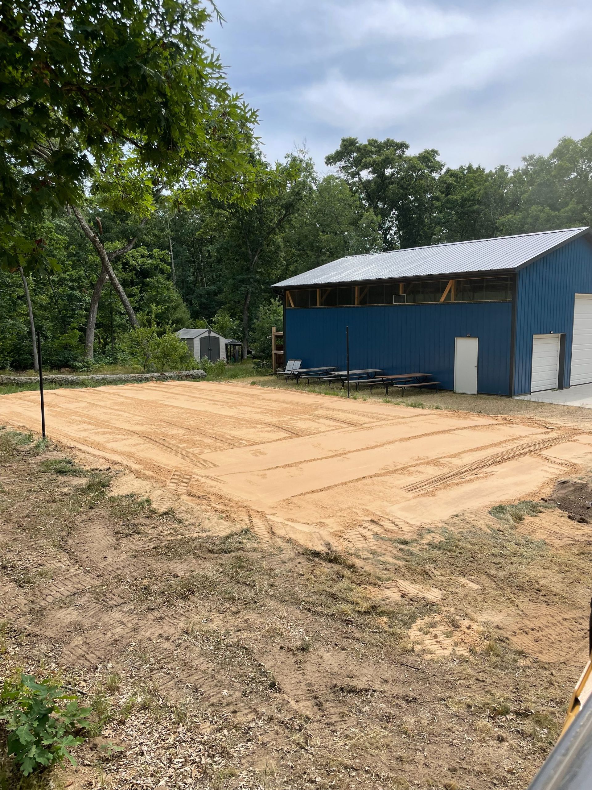 A blue barn stands next to a large, freshly graded rectangular patch of sandy soil surrounded by trees.
