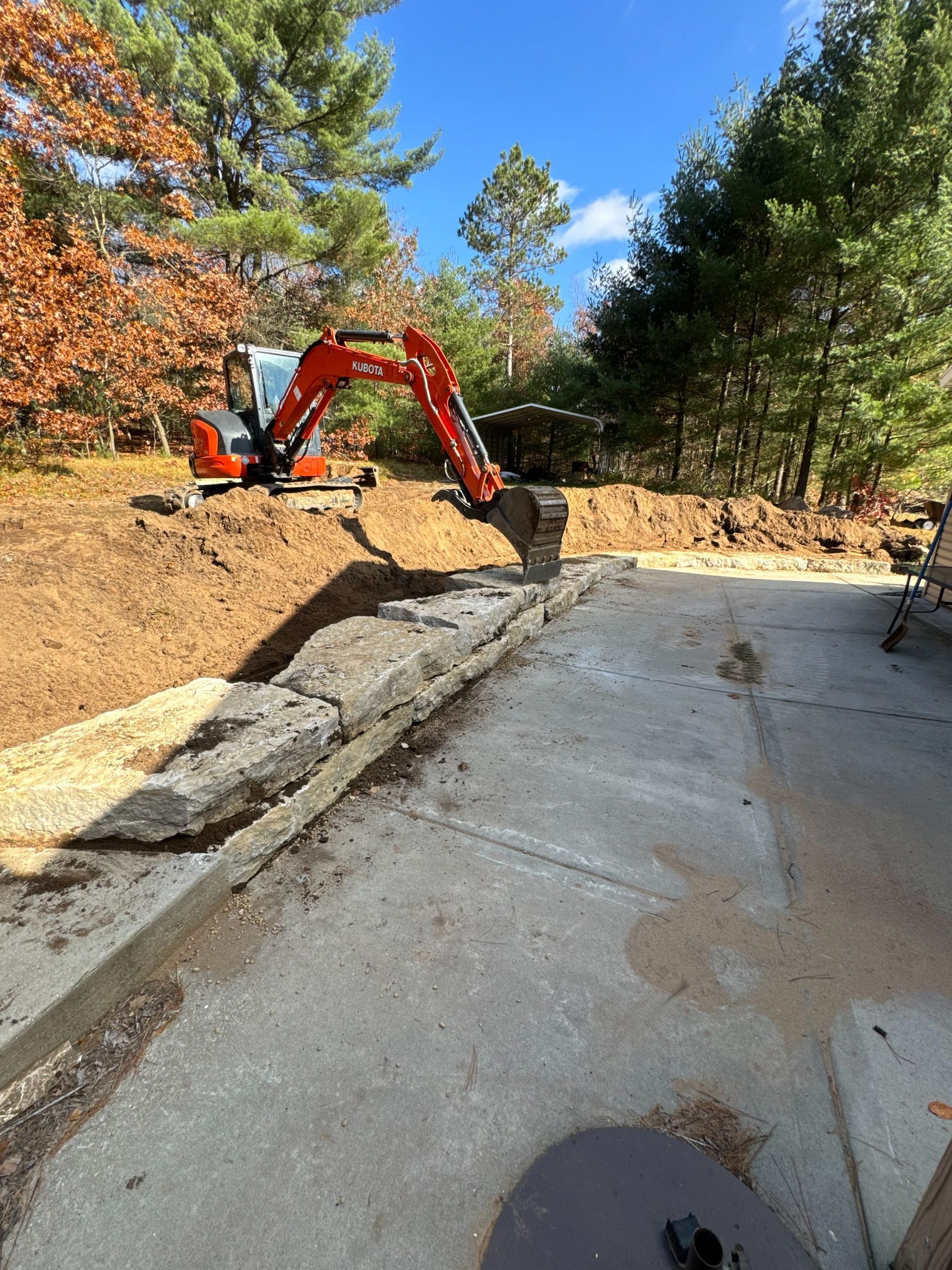 An orange excavator sits on a dirt hill behind a large concrete retaining wall on a sunny day.