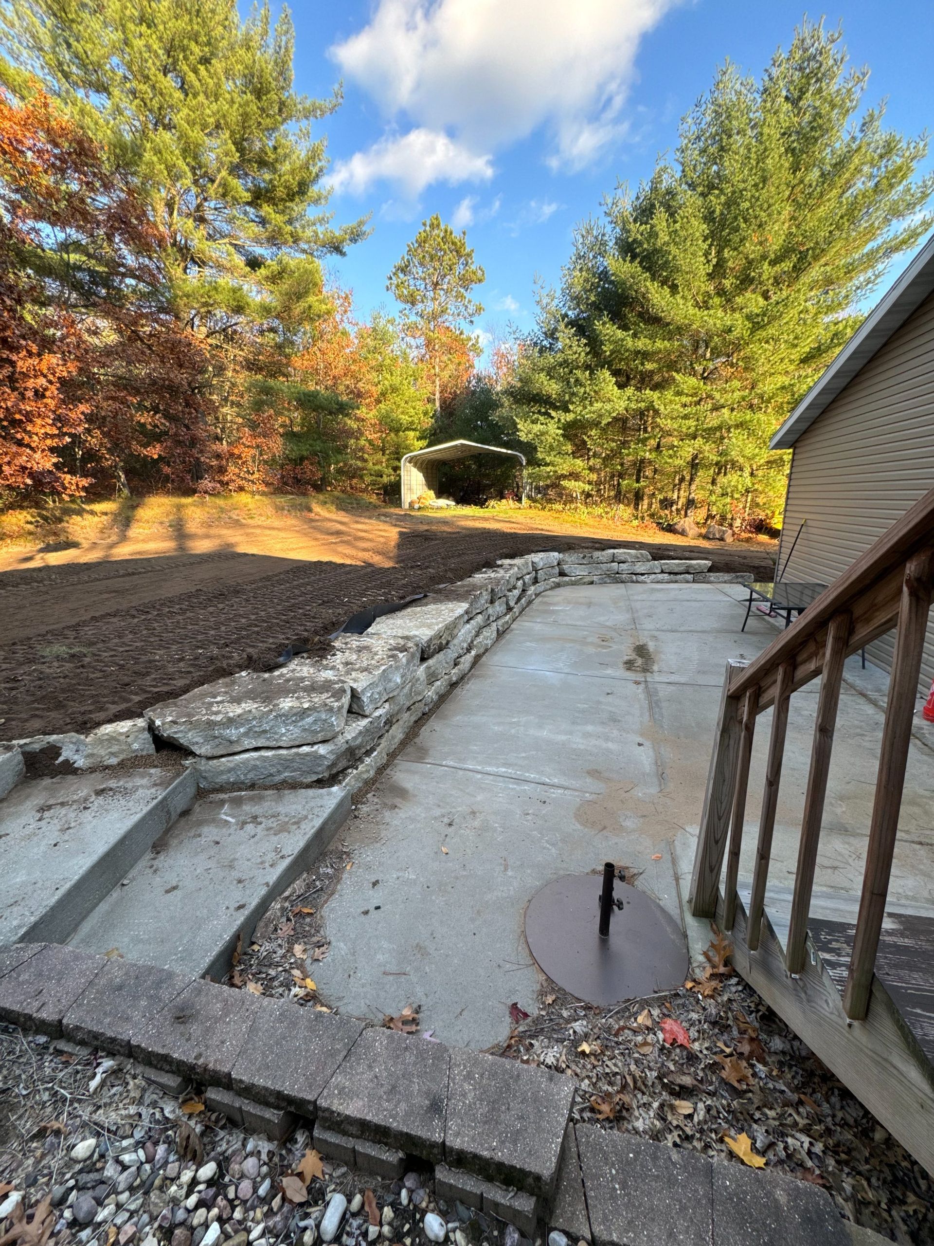 A stone retaining wall bordering a concrete patio, with an area of tilled dirt in the background and a staircase nearby.