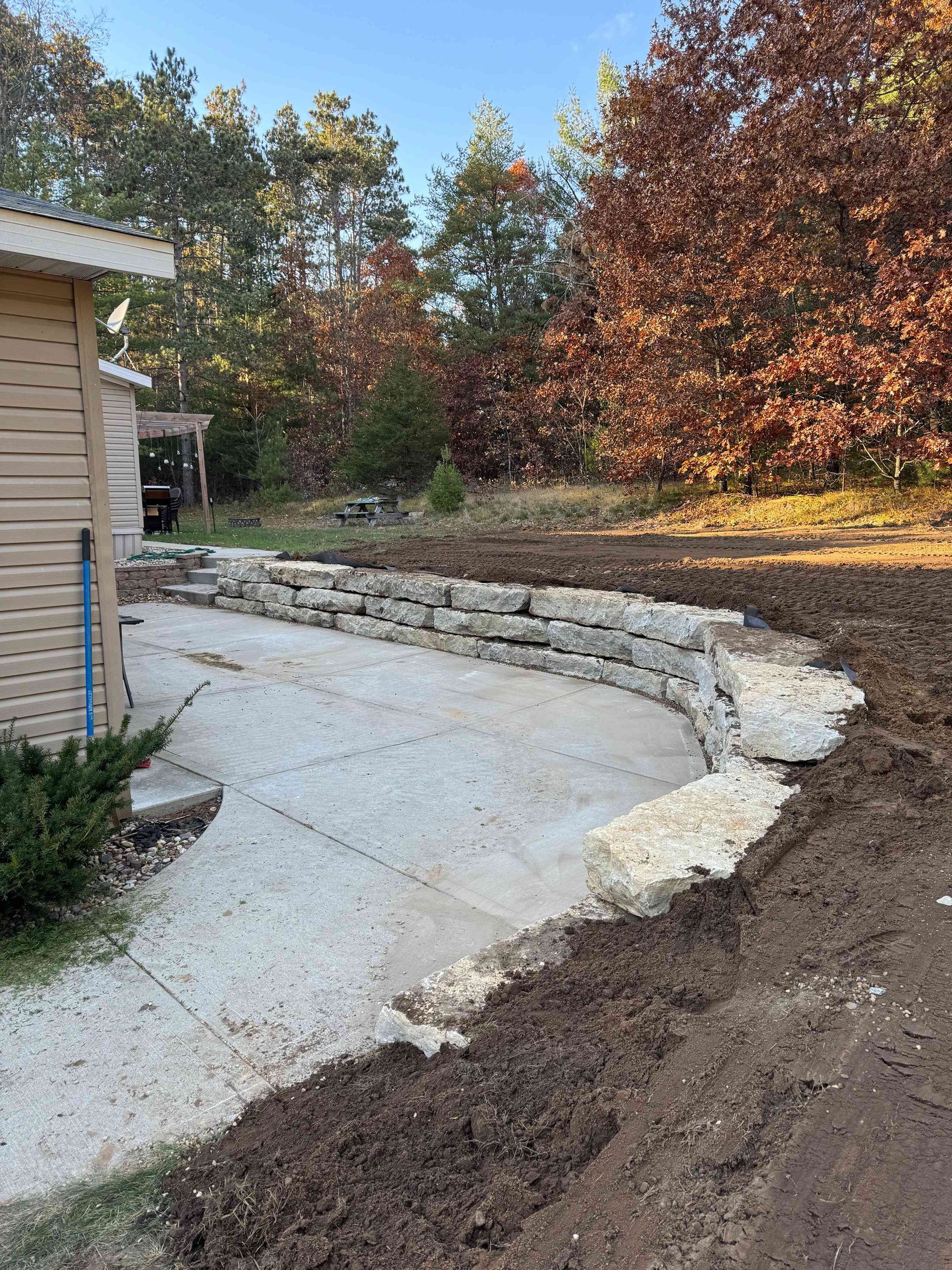 A stone retaining wall bordering a concrete patio, with fresh soil in the foreground and trees in the background.