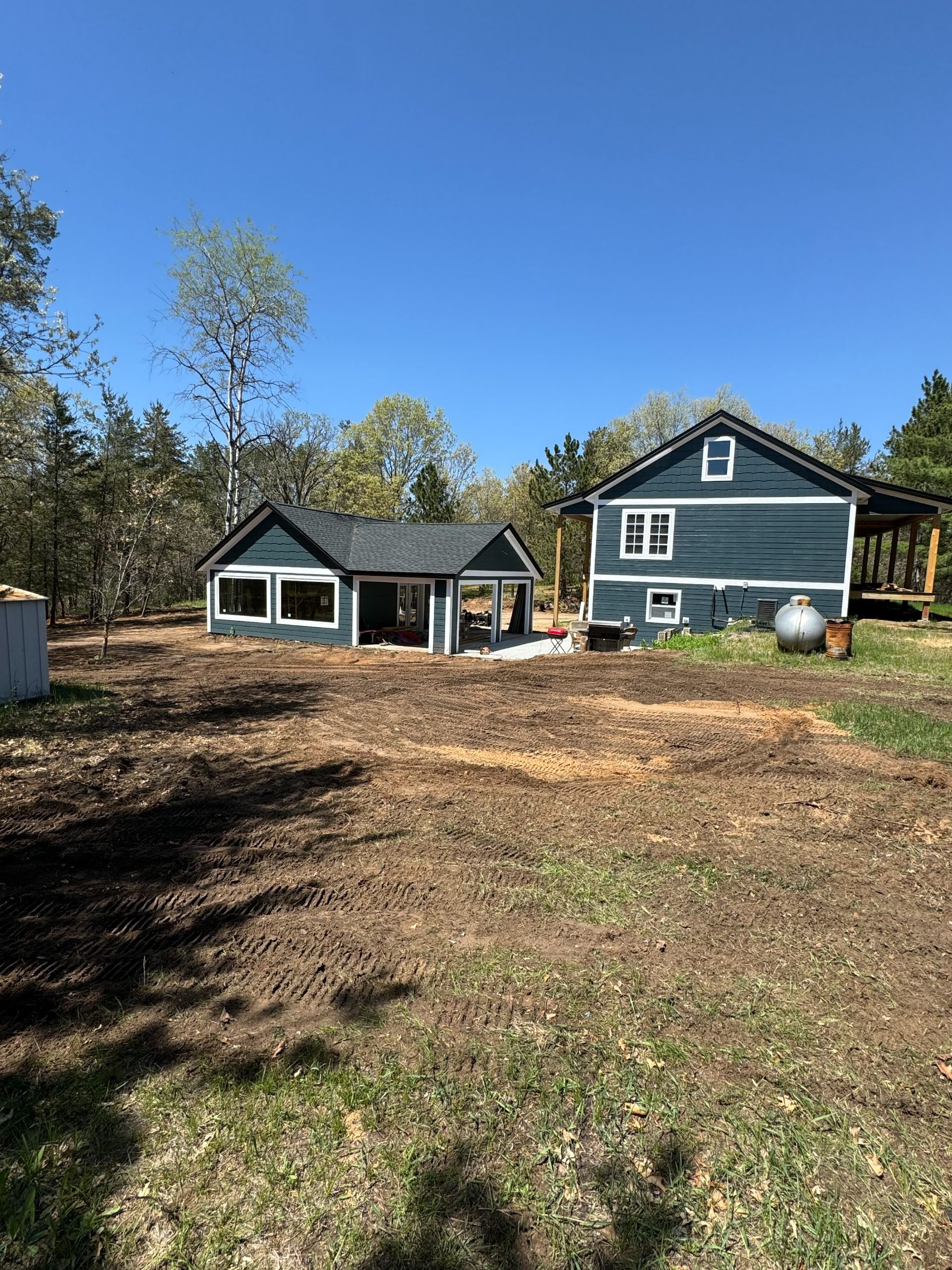Two blue houses with dark gray roofs sit on a large, cleared dirt lot under a clear blue sky.