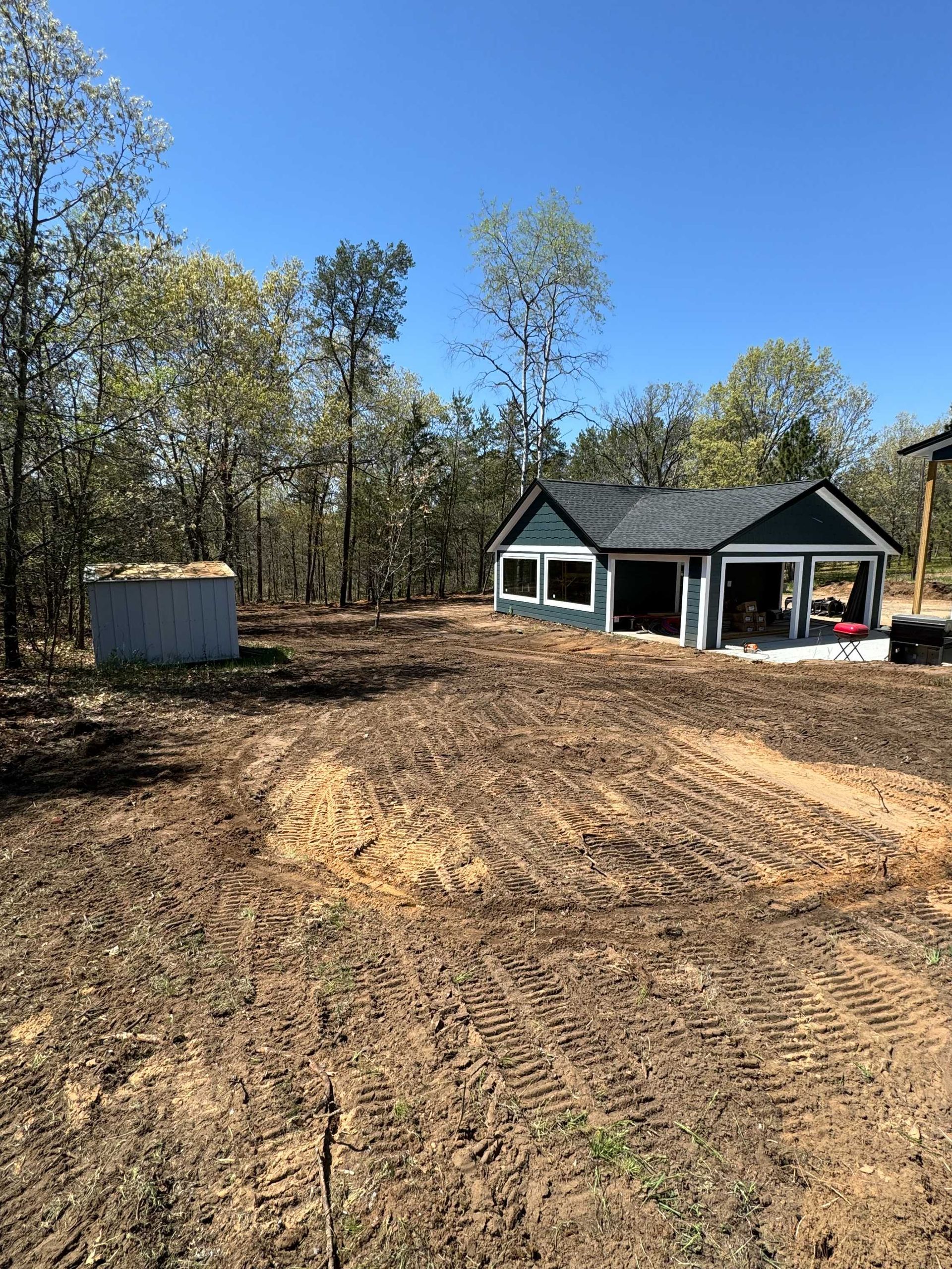 A small, dark-sided building under construction sits on a clearing of tilled earth near a metal shed and a wooded area.