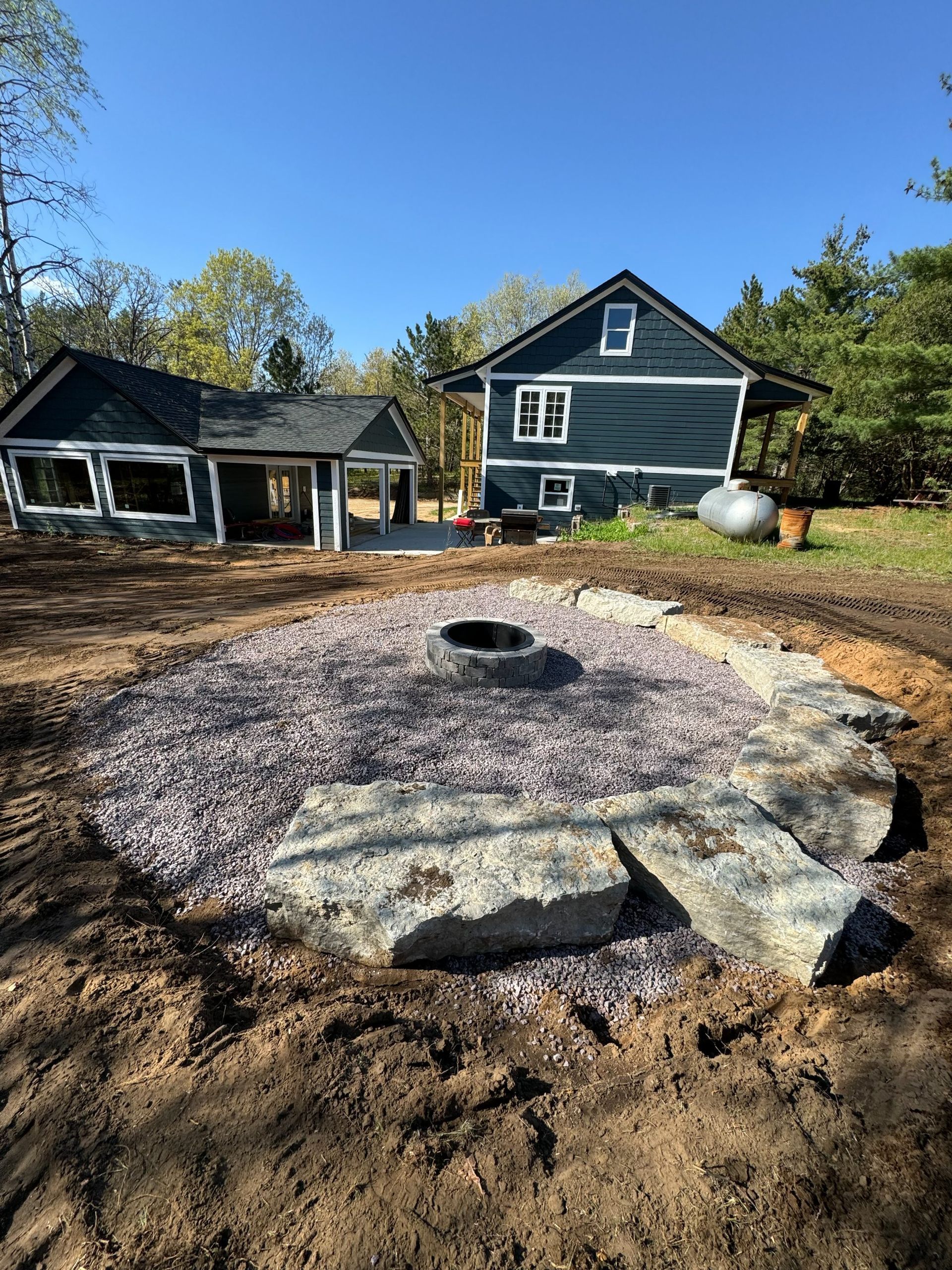 A circular gravel fire pit with a stone border in a dirt yard, in front of two dark-sided houses under a clear blue sky.