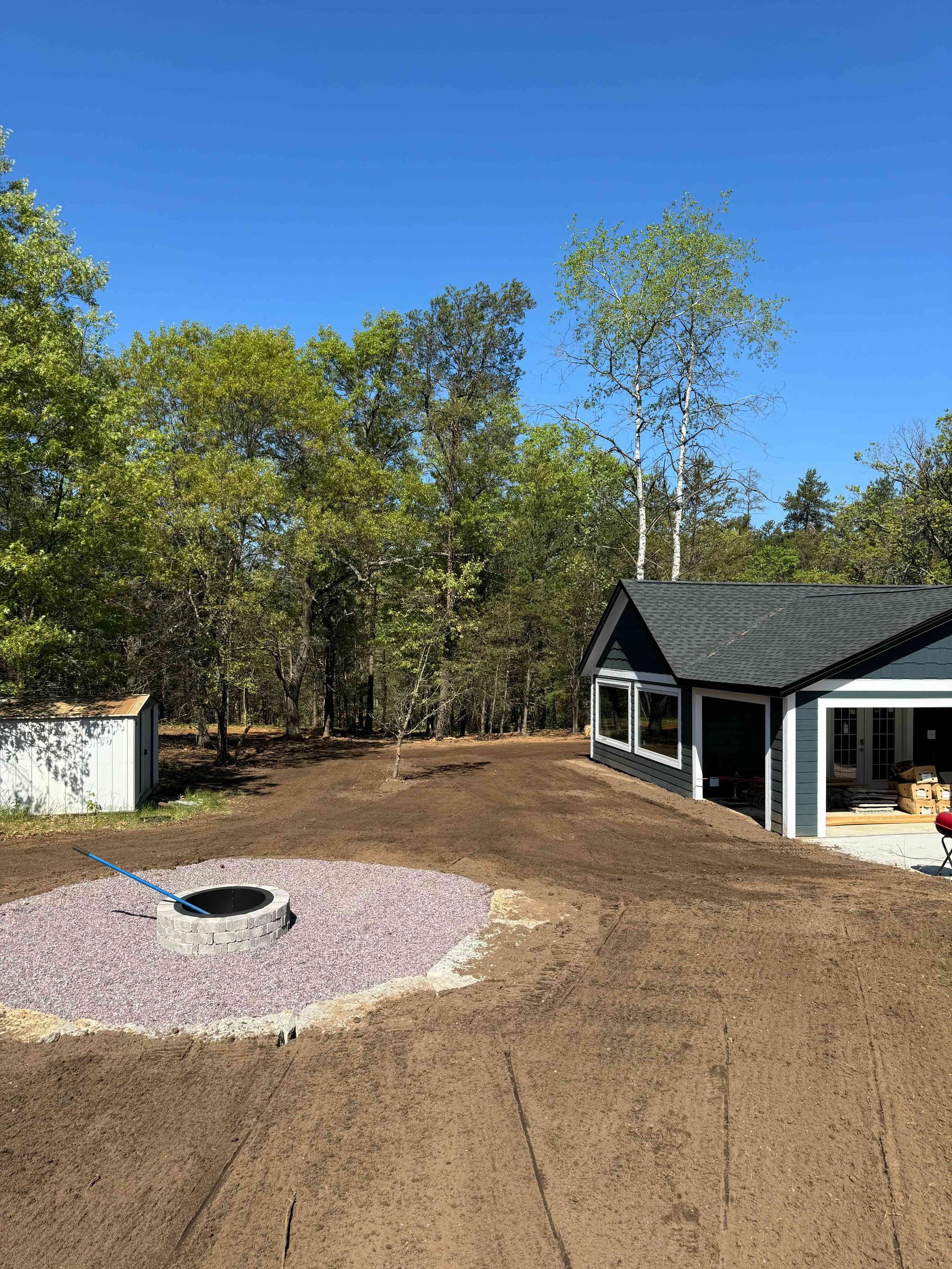 A modern house with gray siding sits beside a fire pit surrounded by purple gravel in a yard of bare dirt and trees.