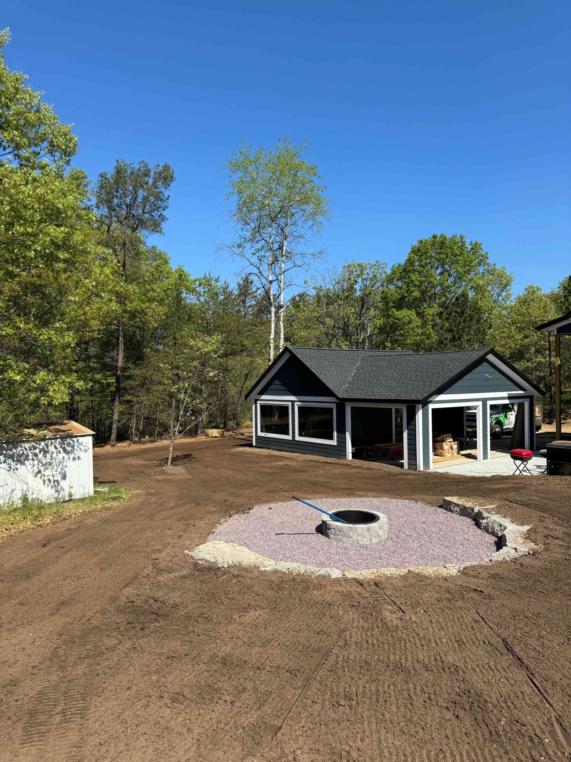 A newly graveled fire pit sits in a dirt yard in front of a blue house under construction with a clear blue sky.