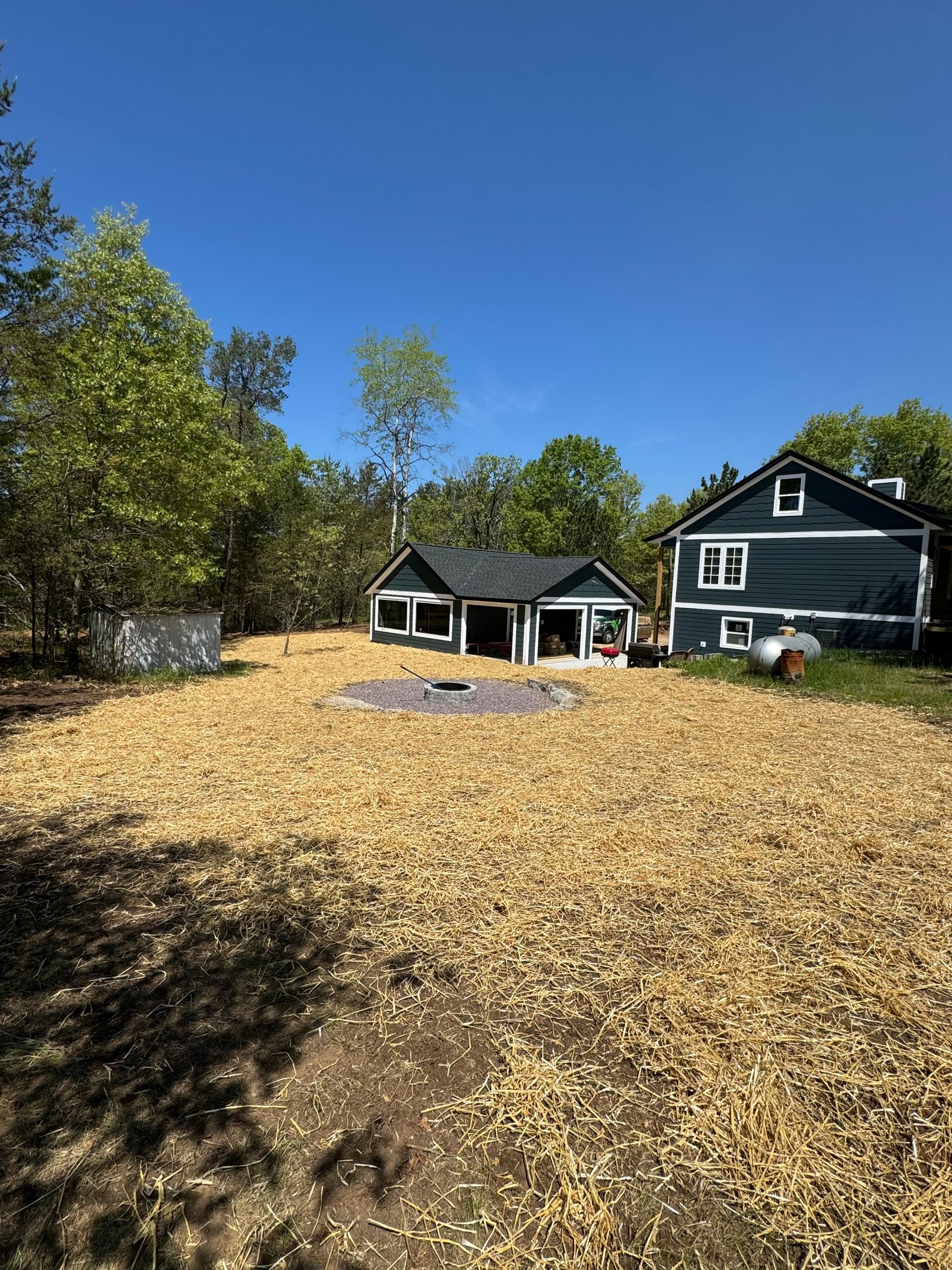 A house and garage sit behind a yard covered in fresh wood chips, with a small circular stone fire pit in the center.