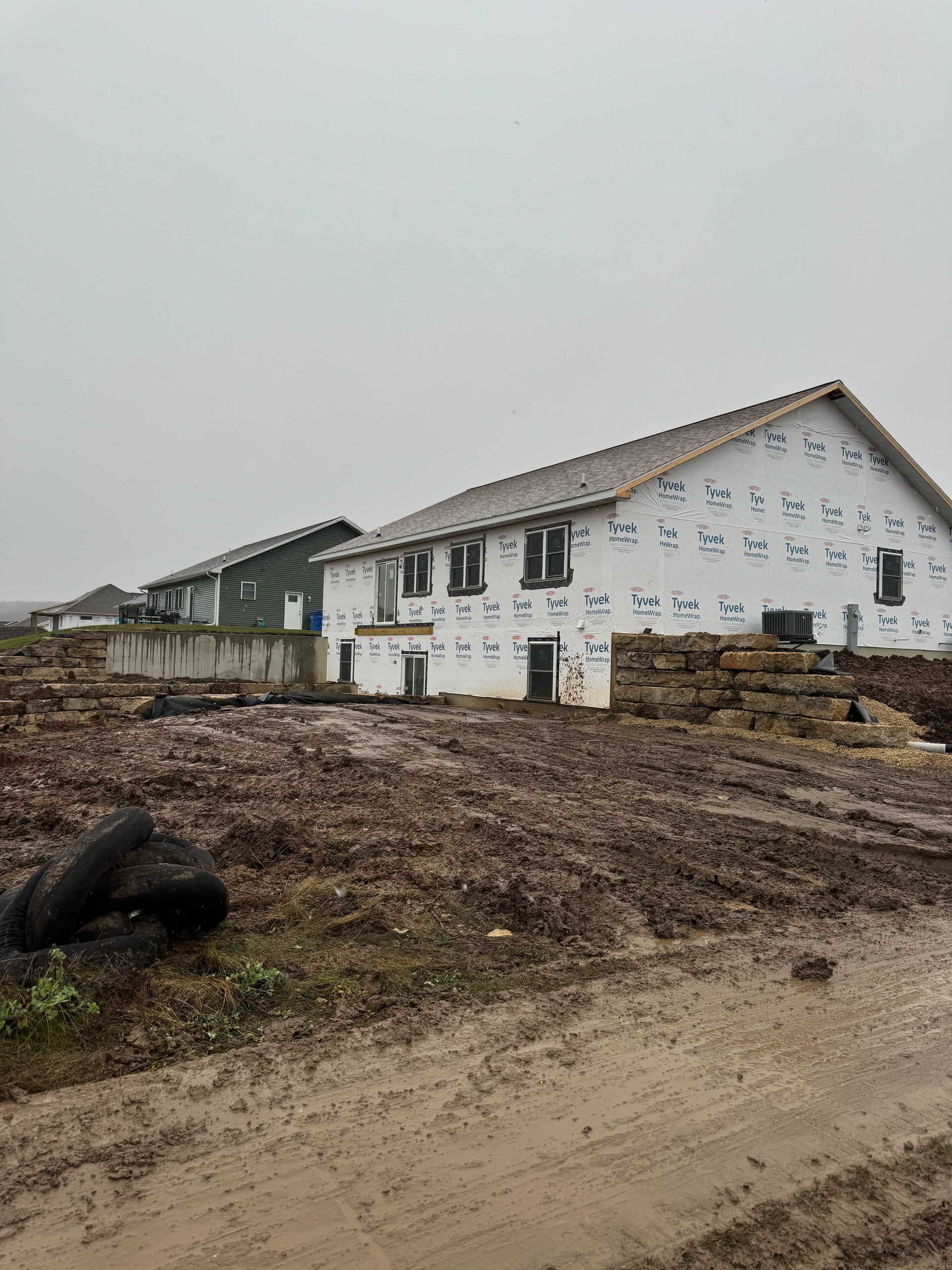 Two unfinished houses with white weather-resistant barriers under construction on a muddy, overcast construction site.