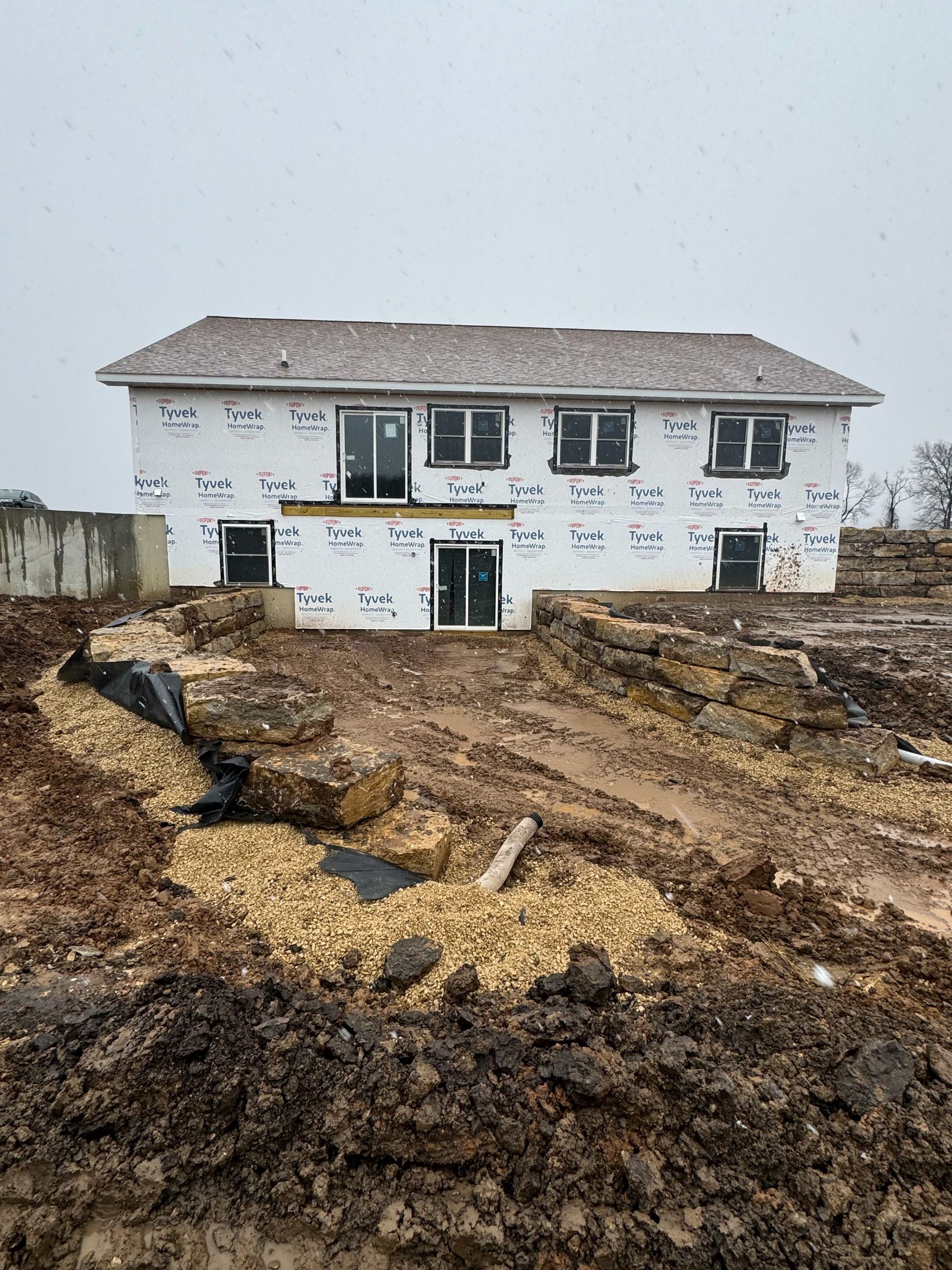 A two-story house under construction with white Tyvek house wrap, windows, and doors, surrounded by dirt and stone walls.