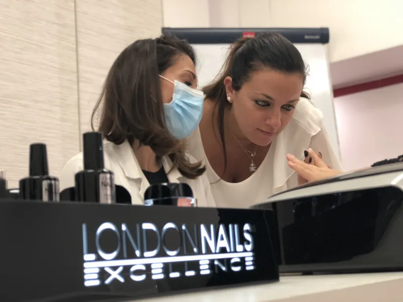 Due donne esaminano una mano in un salone di bellezza, concentrate. Una indossa una mascherina e i prodotti sono etichettati