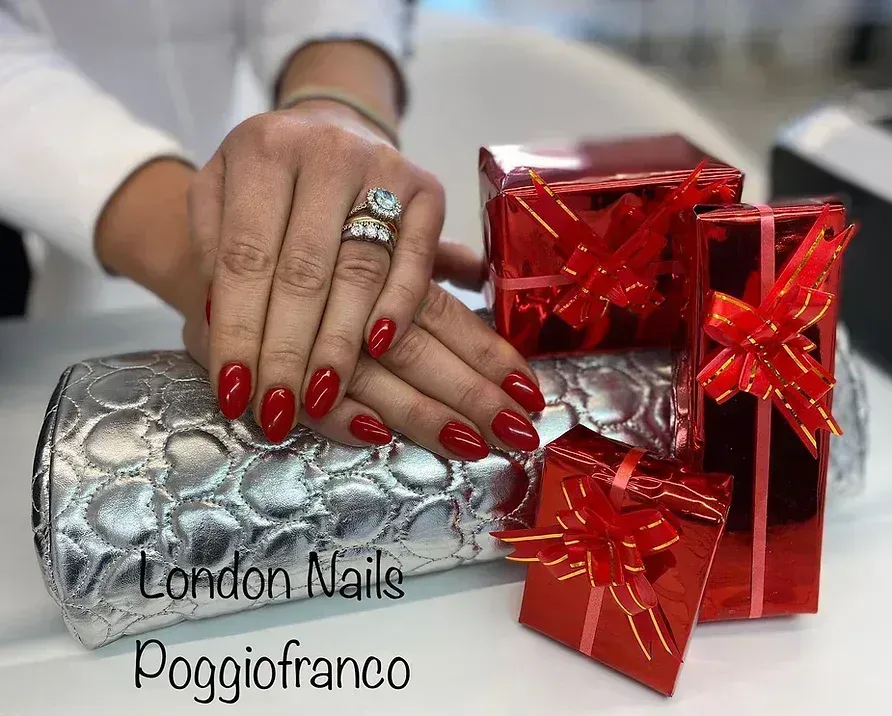 Mani con unghie rosse accanto a pochette argentata e scatole regalo rosse. Testo 
