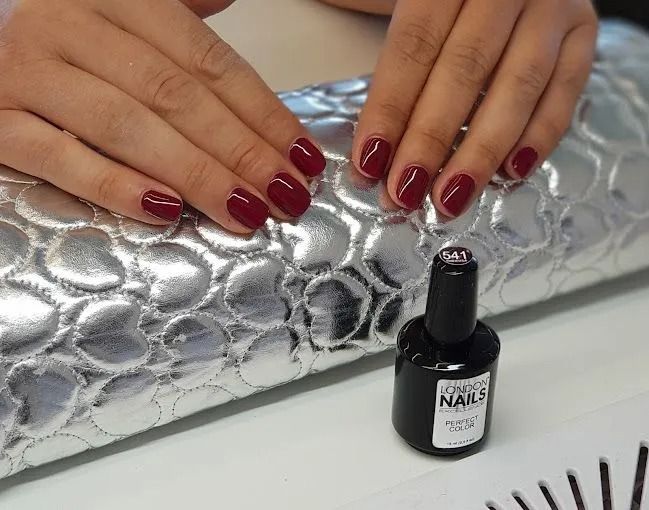 Mani con unghie color borgogna appoggiate su una superficie trapuntata argentata accanto a una boccetta di smalto.