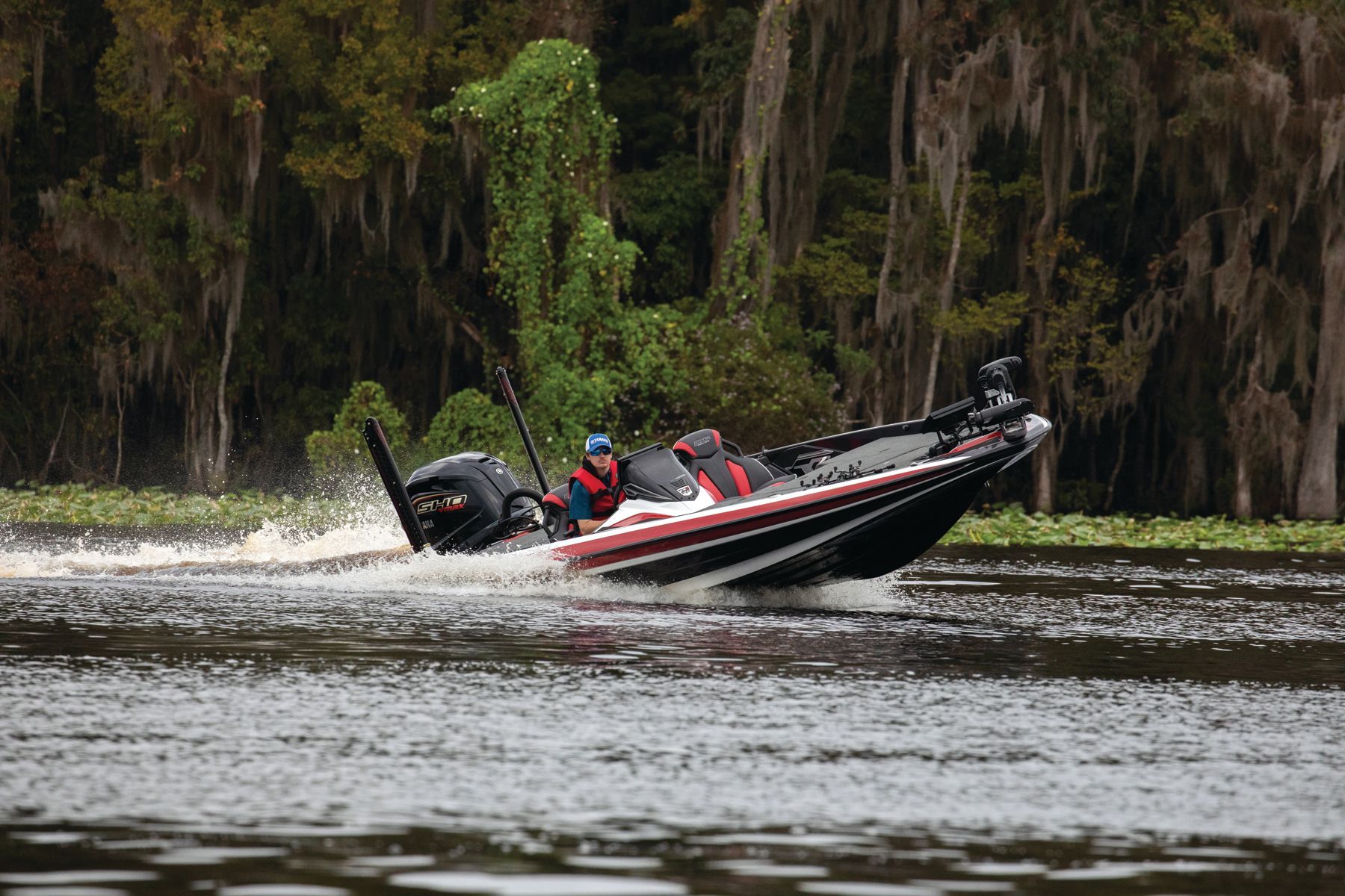 Bass boat speeding on water, person at the helm. Dark boat, splash, green foliage in background.