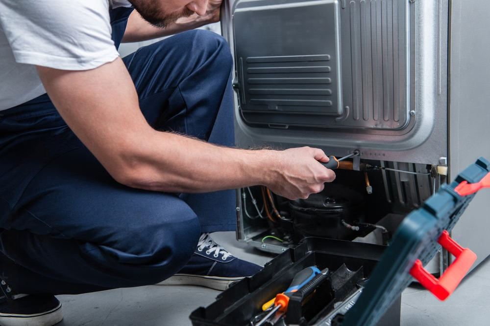 a man is kneeling down fixing a refrigerator with a screwdriver .