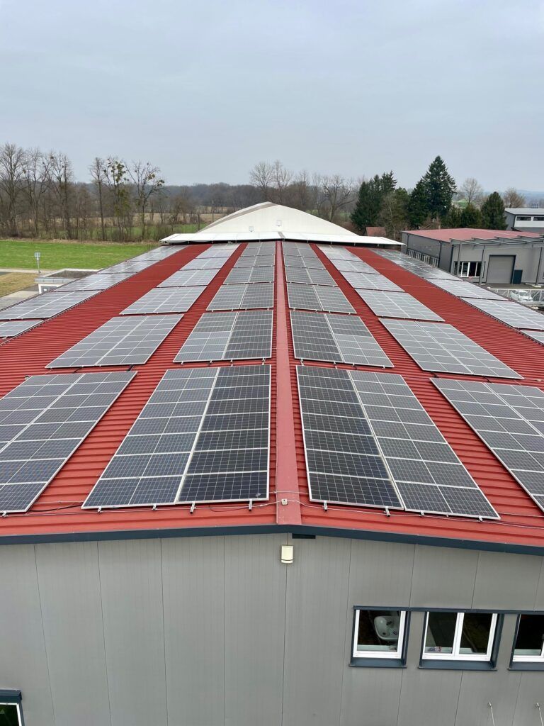 Solarpaneele auf einem roten Lagerhausdach erzeugen erneuerbare Energie. Bewölkter Tag, im Hintergrund Bäume.