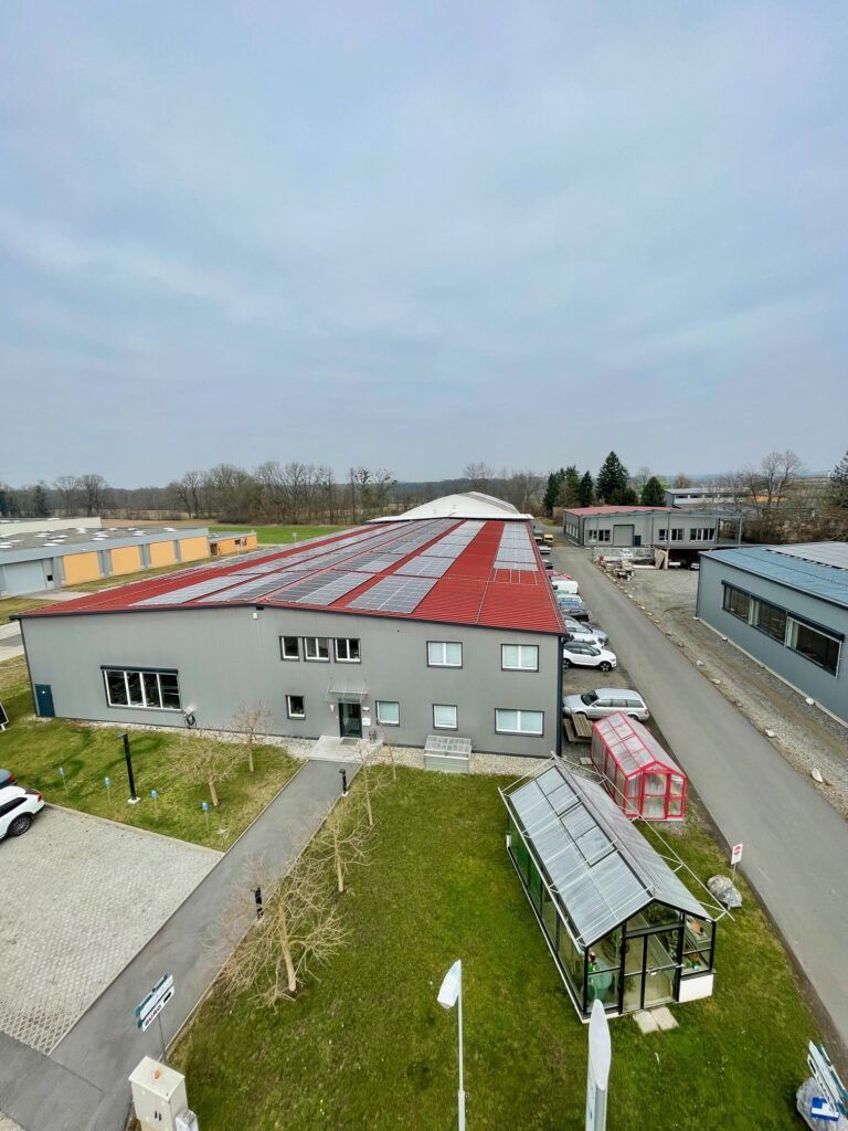 Industriegebäude mit rotem Dach, Solarpaneelen und einem Gewächshaus unter bewölktem Himmel.