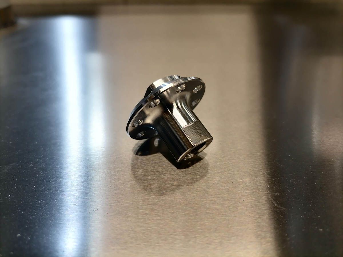 Glänzender Metallknopf mit sechseckiger Basis und kreisförmigem Flansch auf einer Metalloberfläche.