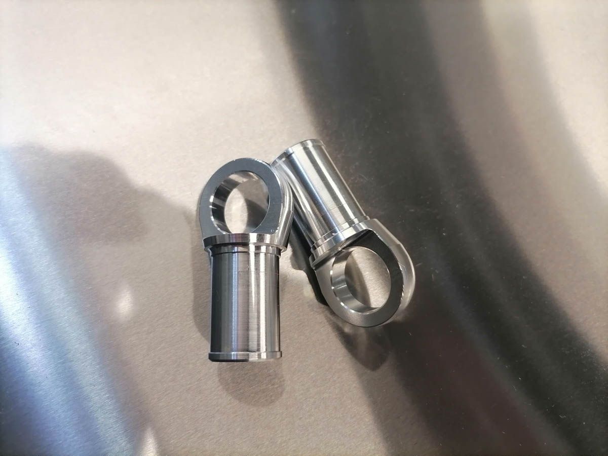 Zwei glänzende Metallstangenendverbinder mit Ösen, die auf einer reflektierenden Oberfläche ruhen.
