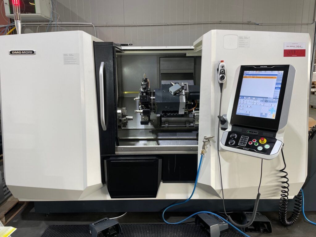 Eine weiße CNC-Maschine mit Bedienfeld und geöffneter Zugangstür in einer Fabrikumgebung.