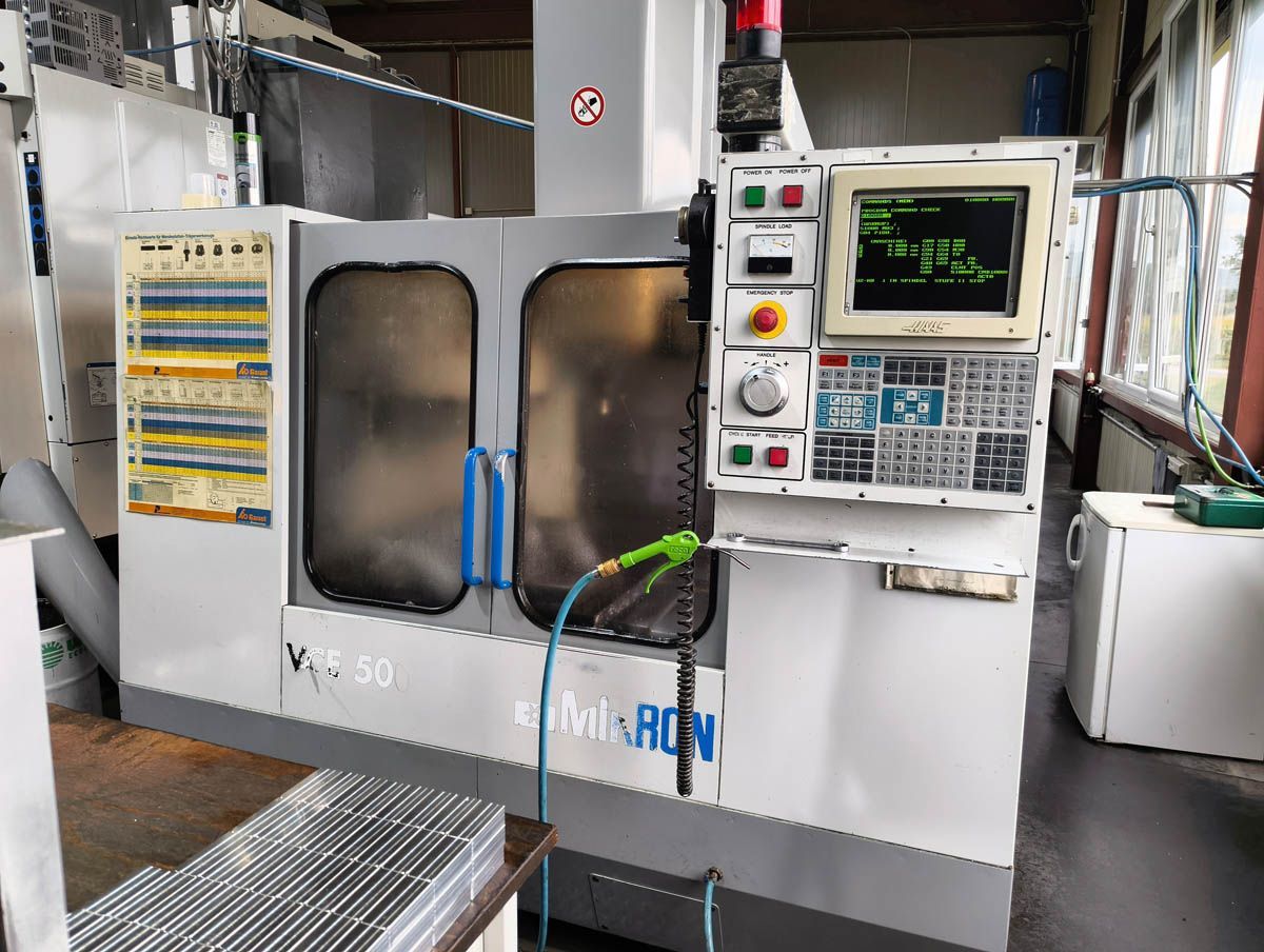 Eine große, graue CNC-Maschine mit Bedienfeld und geschlossenen Türen. Ein Wasserschlauch ragt heraus.