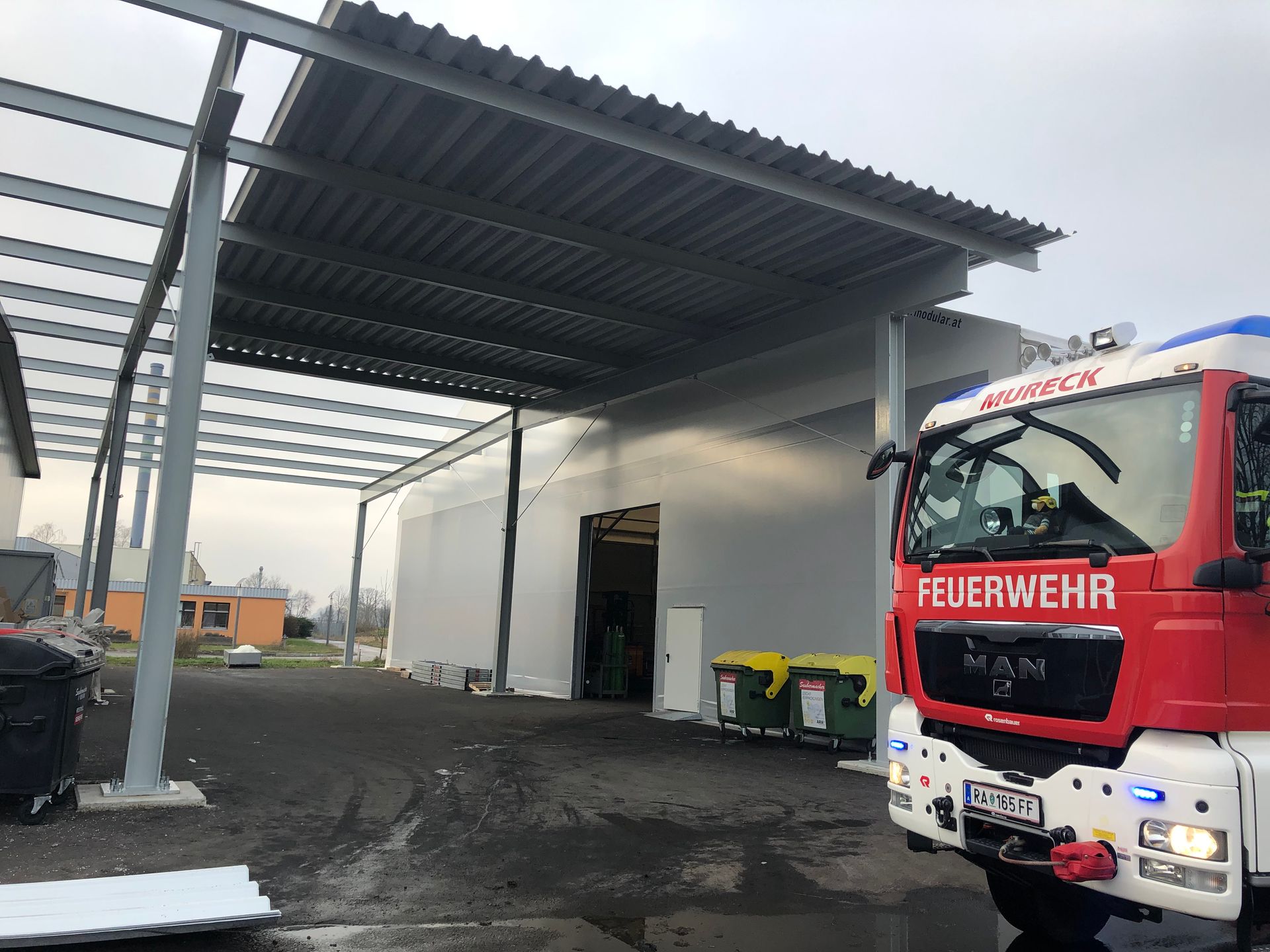 Feuerwehrwagen an einem Gebäude mit offener Fahrzeughalle. Roter Feuerwehrwagen mit der Aufschrift „Feuerwehr“.